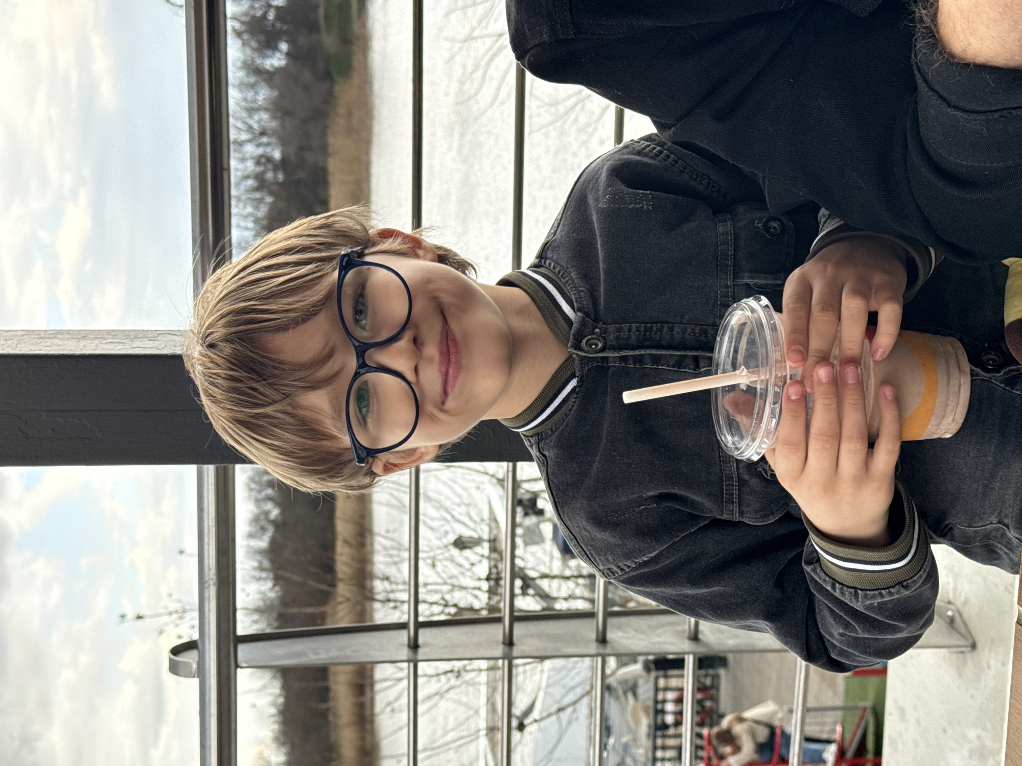 A young child wearing round glasses and a dark denim jacket is holding a plastic cup with a straw. The child is looking at the camera with a slight smile. The background shows a blurred cityscape behind a metal railing. While Rostov-on-Don cannot be confirmed, the photograph may be situated inside a building with a river view.