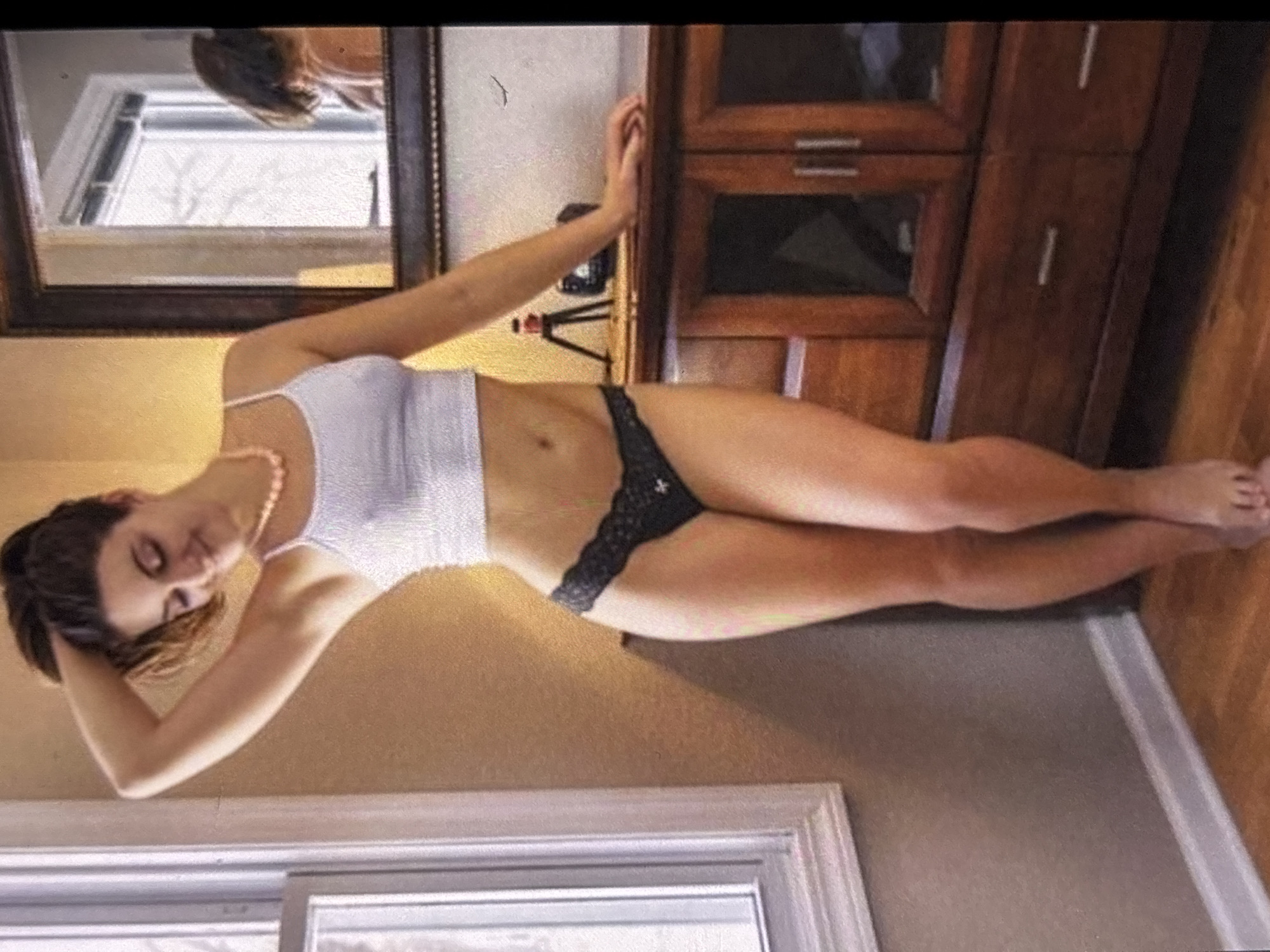 A woman is standing in an indoor setting. She is wearing a white crop top and black lace underwear. She has her hand on her head and is looking down. To her right is a wooden cabinet with a mirror above it. The mirror reflects another woman. The room has a window on the left and hardwood floors. The lighting appears to be natural and warm.