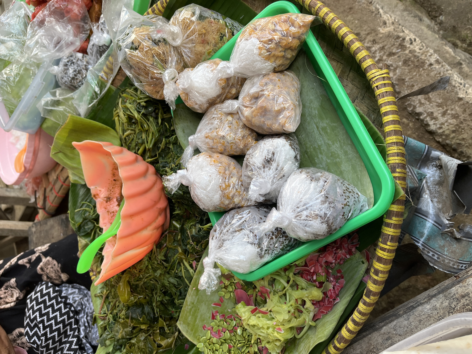 This image captures a close-up, overhead view of a market stall in Tambakreja, Indonesia. The scene is dominated by various food items arranged on banana leaves and in plastic bags. In the foreground, a green plastic tray holds several small, clear plastic bags filled with what appear to be seasoned grains or small fried items, some topped with a white powdery substance. To the left of the tray, there's a portion of a green banana leaf serving as a bed for thinly sliced green vegetables, scattered with small green seeds and pinkish flower petals. Behind this, a large pile of cooked green leafy vegetables is visible, with a bright orange, ribbed plastic bowl containing a small green utensil resting on top. In the background, more food items in plastic bags are visible, including some that appear to be dried or fried. The stall appears to be outdoors, with the suggestion of a woven basket base for the display and glimpses of a person's black and white patterned clothing at the top left. The overall impression is of a vibrant, local food market offering fresh produce and prepared snacks.