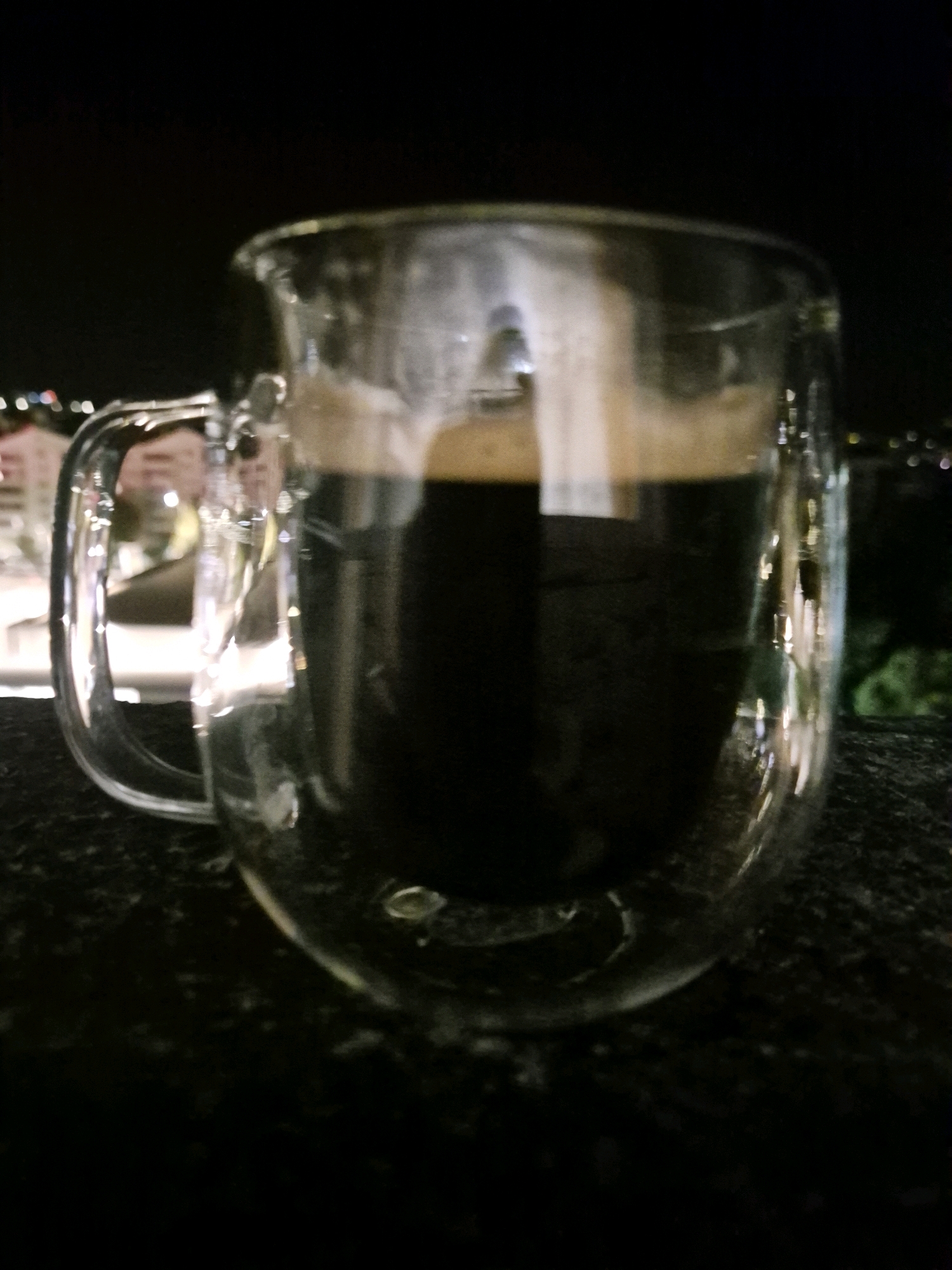 A double-walled glass mug filled with dark liquid, likely coffee or an espresso drink, sits on a dark, textured surface. The mug has a clear glass handle and a visible crema on top of the beverage. The background is dark and out of focus, with blurred lights suggesting an urban or outdoor evening setting. The image is shot from a slightly low angle, with the mug being the primary subject.