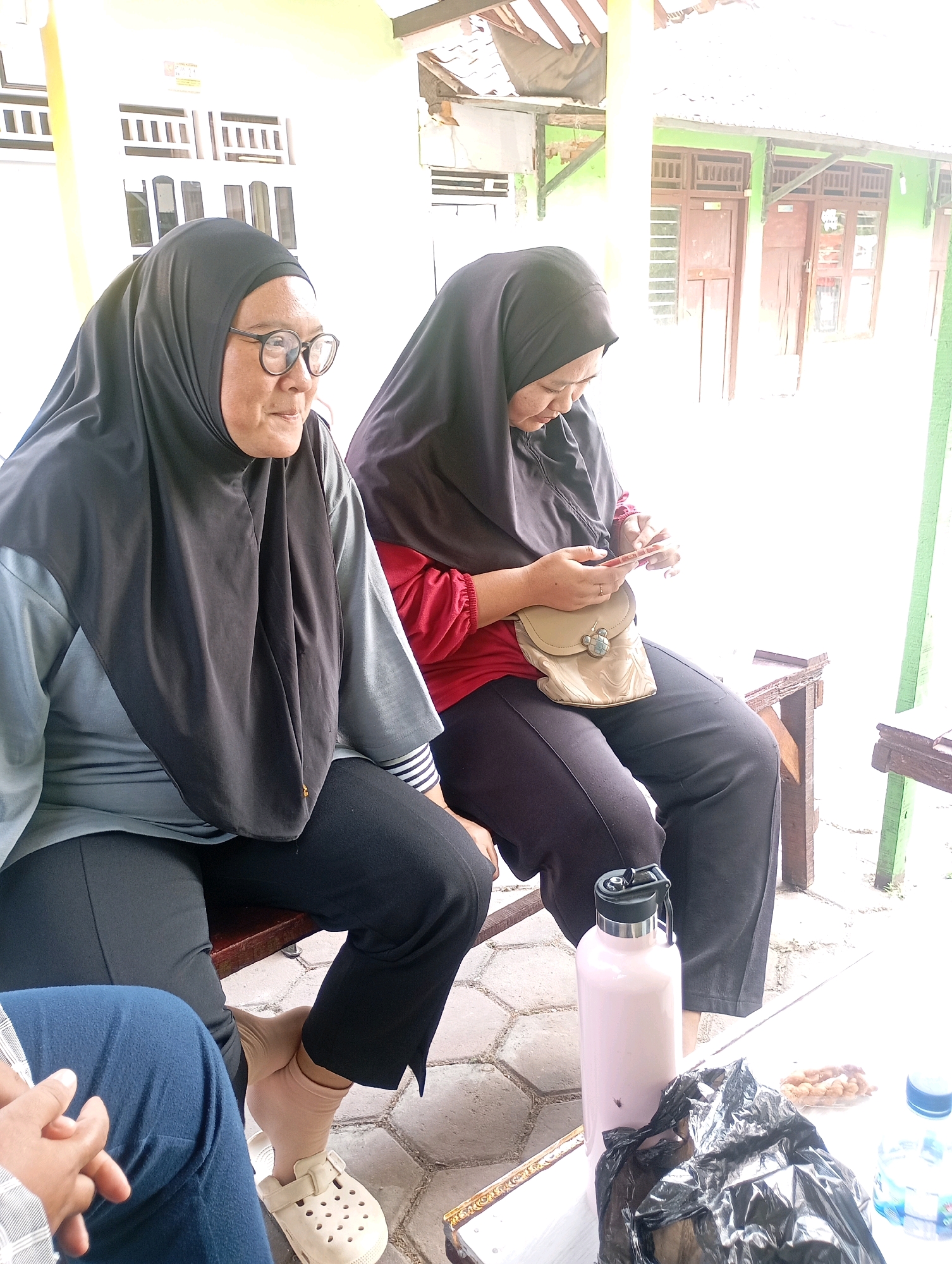 Two women are sitting on a wooden bench outdoors in what appears to be a public area or the grounds of a building in Babelan, Indonesia. The woman on the left, wearing a black hijab and glasses, is looking to her right. She is dressed in a grey and black long-sleeved shirt and black pants. The woman on the right, also wearing a black hijab, is looking down at her phone. She is wearing a red long-sleeved shirt and black pants, and has a small cream-colored crossbody bag with a bear-shaped clasp. In front of them, on a white table, is a light pink water bottle with a black lid, a crumpled black plastic bag, a small pile of snacks, and a partially visible bottle of water. The ground is paved with light-colored stones. In the background, there is a building with green walls and wooden doors and windows, and a wooden roof structure. The overall impression is a casual, outdoor gathering.