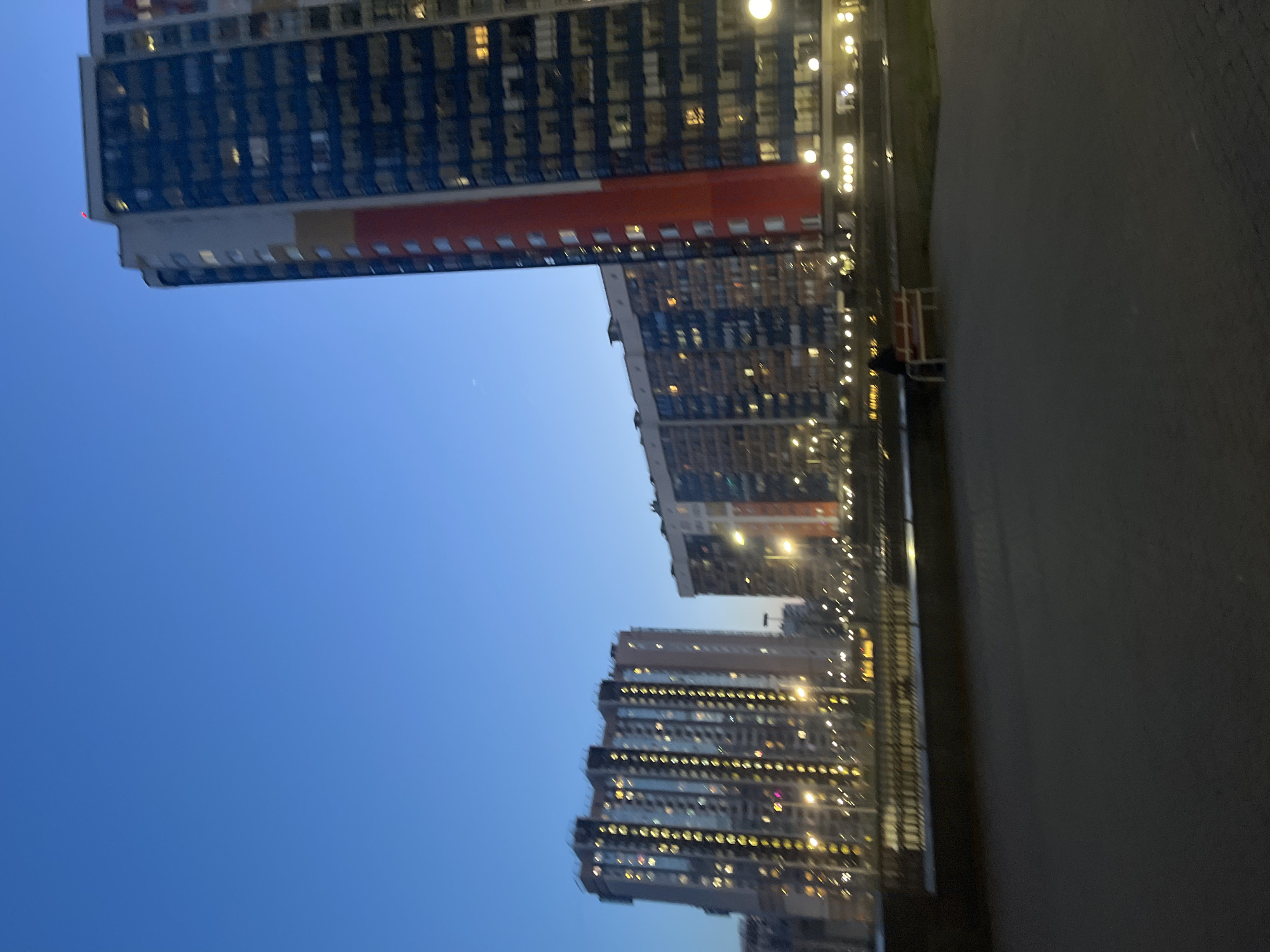 The image shows a view of several tall apartment buildings at dusk in Vsevolozhsk, Russia. The buildings have many lit windows. The sky is blue, and the ground is a dark pavement. A street light is visible. There are no people visible.