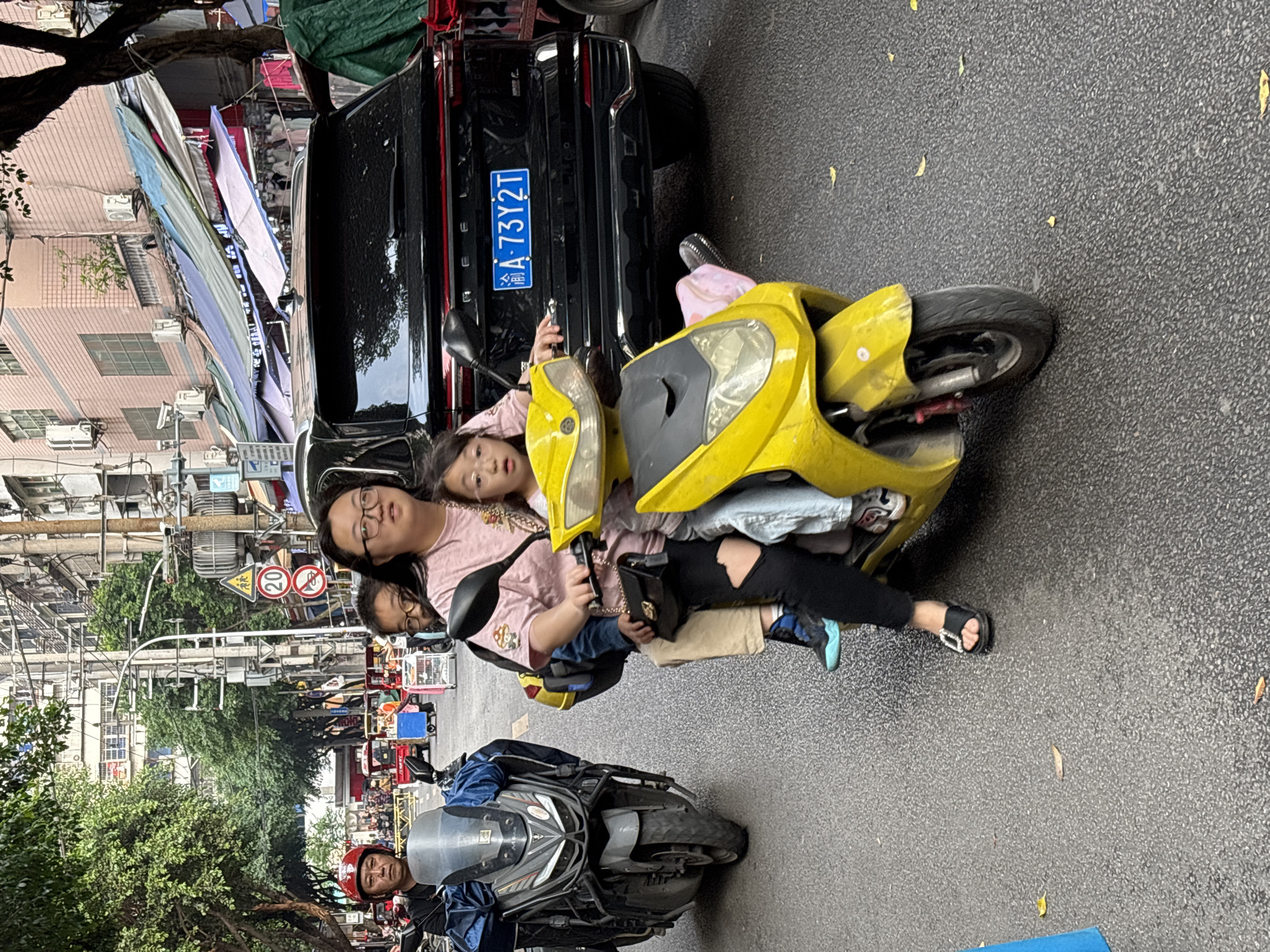 The image shows a woman and two children on a yellow scooter parked near a black vehicle with the license plate 渝A·73Y2T. The vehicle appears to be a SUV. The woman is wearing glasses and a pink shirt. One child is sitting in front of her on the scooter, while the other is seated behind. A black purse is hanging from the handlebar. A man on another scooter, wearing a red helmet, is positioned at the bottom of the frame, facing the camera. The scene is on a street in Jiangjin, China, where there are shops and food stalls visible in the background, with awnings extended. Traffic signs can also be observed further down the street.