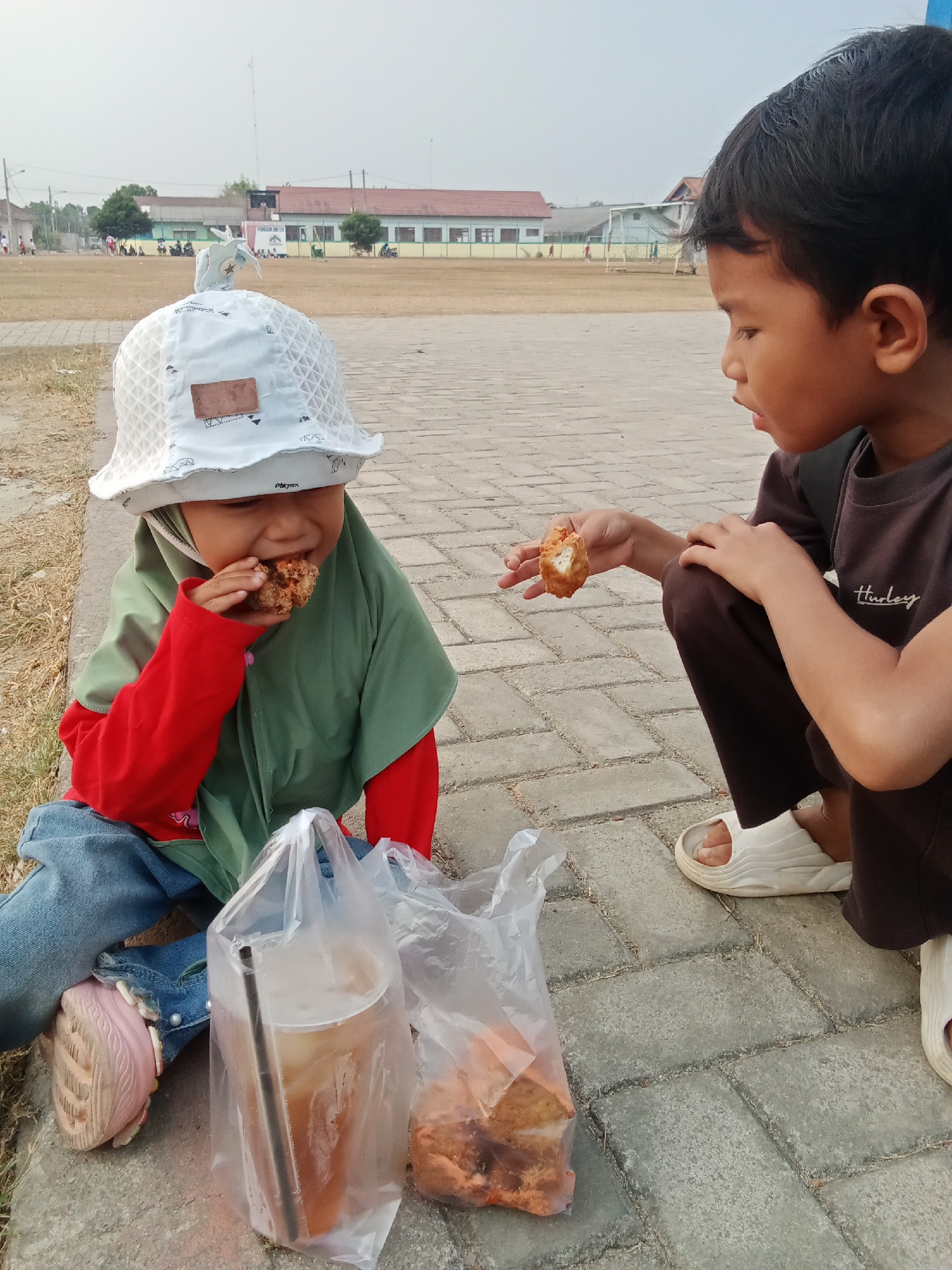 Two young children are sitting on a paved surface outdoors. On the left, a child wearing a white patterned hat, a green hijab, a red long-sleeved shirt, and blue jeans is eating a piece of fried food. To the right, a child wearing a dark brown t-shirt and dark brown pants, and white slip-on sandals, is holding a piece of fried food and looking at the other child. In front of the children are two plastic bags containing drinks and fried food. The background shows a dry, grassy field with buildings and trees, consistent with a schoolyard or park in Toto Katon, Indonesia. The sky is hazy and overcast.