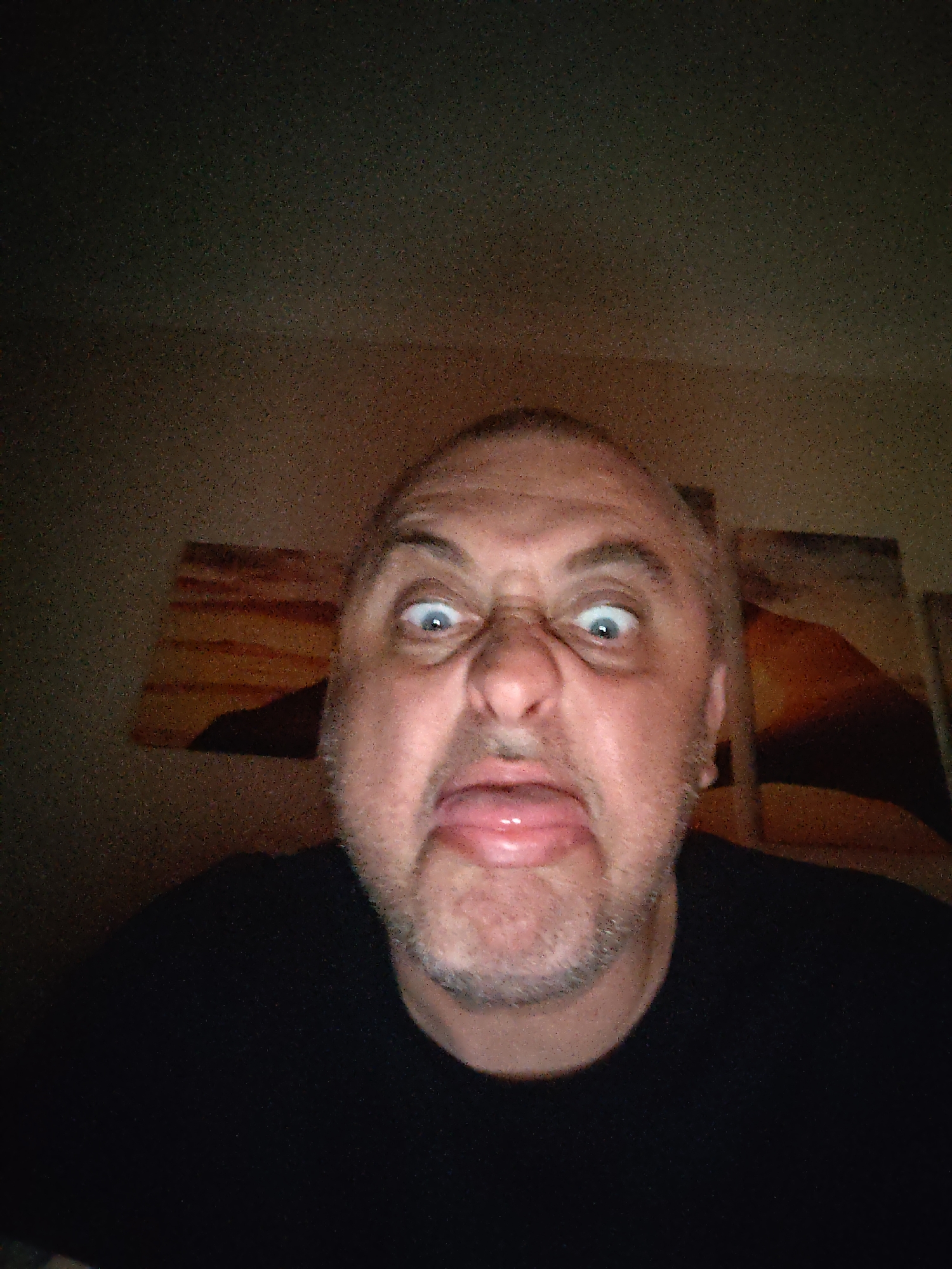A close-up, low-light photograph captures a light-skinned, middle-aged man making a humorous facial expression. He has short hair and a neatly trimmed beard or stubble. His eyes are wide open and slightly crossed, his nose is scrunched, and his tongue is extended outwards from his mouth. He is wearing a black t-shirt or top.
The setting appears to be indoors, with a dimly lit, textured wall in the background, possibly corkboard. Two abstract or landscape-style artworks, partially obscured by shadow and the man's head, are visible on the wall behind him. The low lighting conditions result in a noticeable amount of image grain. The overall emotion conveyed is playful and silly. There is no visible text in the image. The location is contextualized as Huntingdonshire, United Kingdom.
