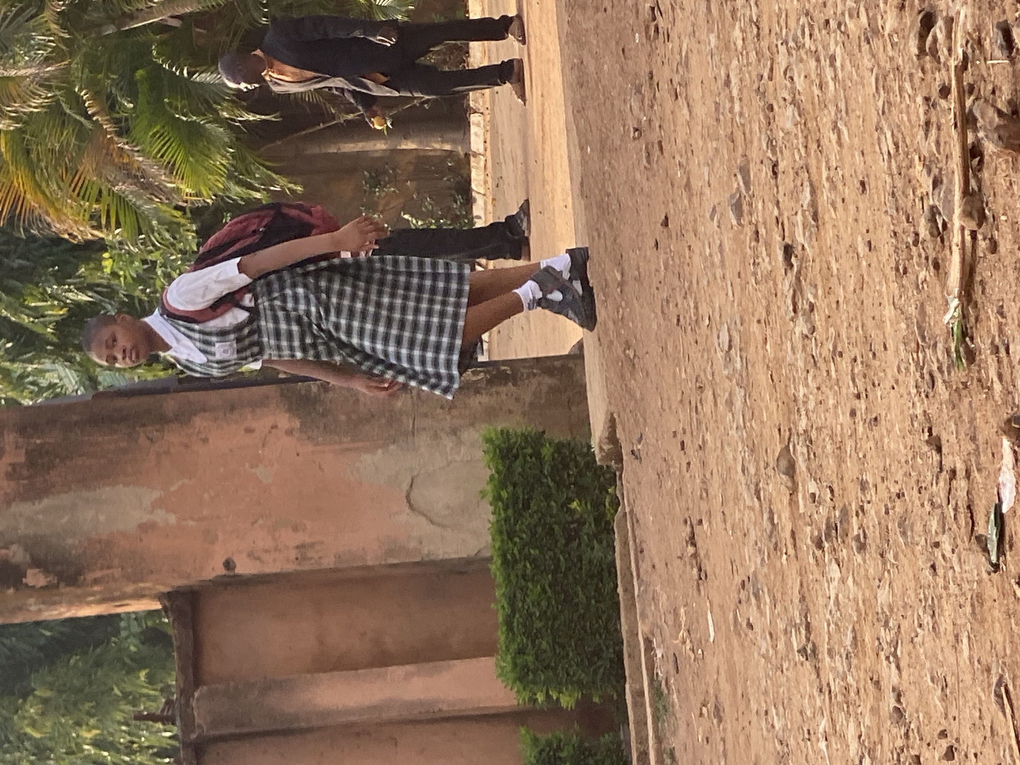 The image captures an outdoor scene in what appears to be Maigizo, Nigeria, featuring a young person in a school uniform as the primary subject. The setting includes a dusty, light brown ground or path, with some worn, light orange-brown architectural structures forming a wall or building in the mid-ground. Lush green, palm-like foliage and other green shrubs are visible in the background, suggesting a warm climate.
The central figure is a young person, likely a female student, standing on the dirt path, looking directly towards the camera. She is wearing a white, short-sleeved school shirt with a dark-colored, possibly maroon, backpack slung over her shoulders. A small, unreadable white badge is pinned to the left side of her shirt. She wears a plaid (tartan) skirt in shades of green and white, white ankle socks, and dark-colored sneakers with white soles. Her posture is relaxed, with her arms gently bent in front of her.
In the further background, partially obscured by the foliage, another person (an adult male) is seen standing or walking. He is wearing dark trousers and a light-colored top. The lighting is bright and sunny, indicating clear weather, likely mid-day or late morning/early afternoon, given the strong, defined shadows. There is no discernible text other than the blurry badge on the student's shirt. The scene depicts a mundane, everyday moment, possibly near a school or on the way to or from one.