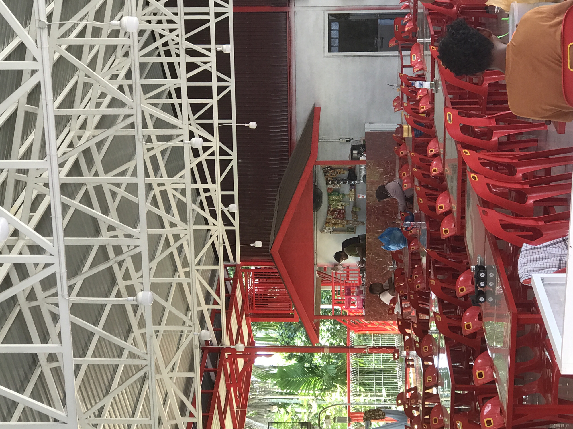 A casual, open-air restaurant scene in Bulo Gogo, Indonesia. The restaurant features a white, lattice-structured roof and red accents throughout. Rows of red plastic chairs are stacked at tables, and several individuals are seated, appearing to be patrons or staff. A bar area displays stocked shelves with food items. Outside, lush greenery is visible through the open walls of the restaurant.