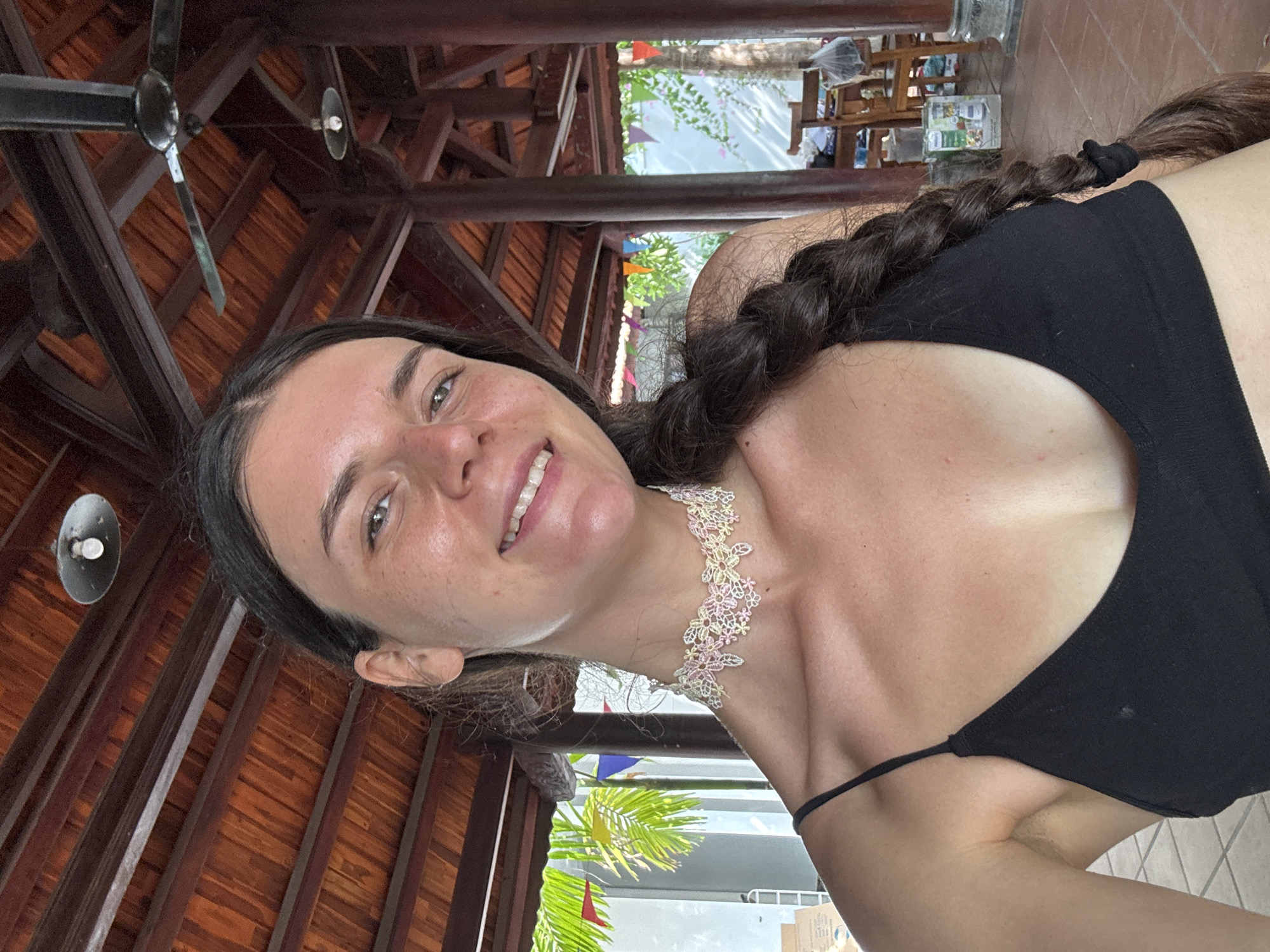 A young woman with dark hair in a braid is pictured from the chest up. She is wearing a black tank top and a light-colored lace choker with floral detailing. She is smiling, and her skin has a light tan with freckles. The background appears to be an open-air structure with a wooden roof and ceiling fan. Palm fronds and colorful flags are visible in the distance, suggesting a tropical location. The floor is tiled with reddish-brown tiles. The overall ambiance is casual and relaxed, consistent with a vacation setting in Mũi Né, Vietnam.