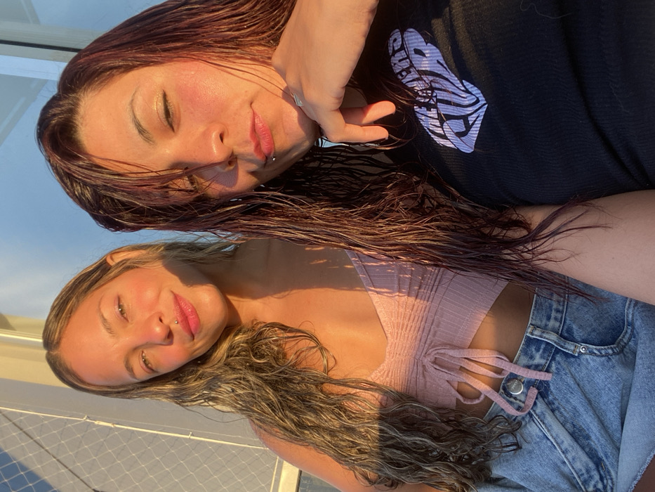Two women pose closely together. The top woman has dark, wet hair, fair skin, and a lip piercing. She wears a black shirt with a graphic design. The bottom woman has long, wavy, highlighted hair, fair skin, and a lip piercing. She wears a light pink crop top and denim shorts. The picture is taken at an angle and bathed in golden sunlight. The background is indistinct, possibly a rooftop or balcony.