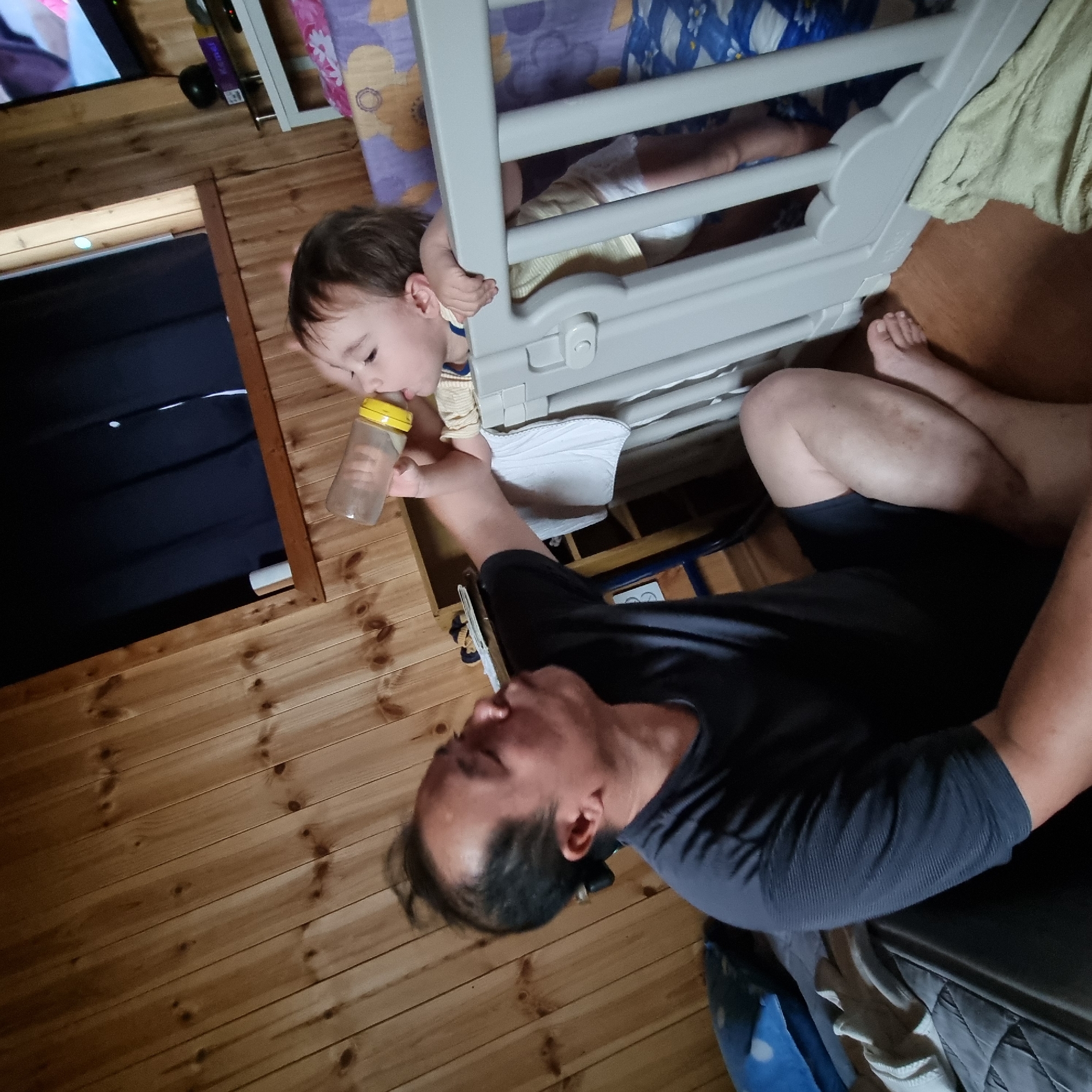 In Dunnae-myeon, South Korea, a man is feeding a baby from a bottle. The man is seated on the floor, leaning towards a baby who is in a crib. The baby is holding the bottle and drinking from it. The room has wood paneled walls and a television is visible in the background. The lighting in the room is dim.