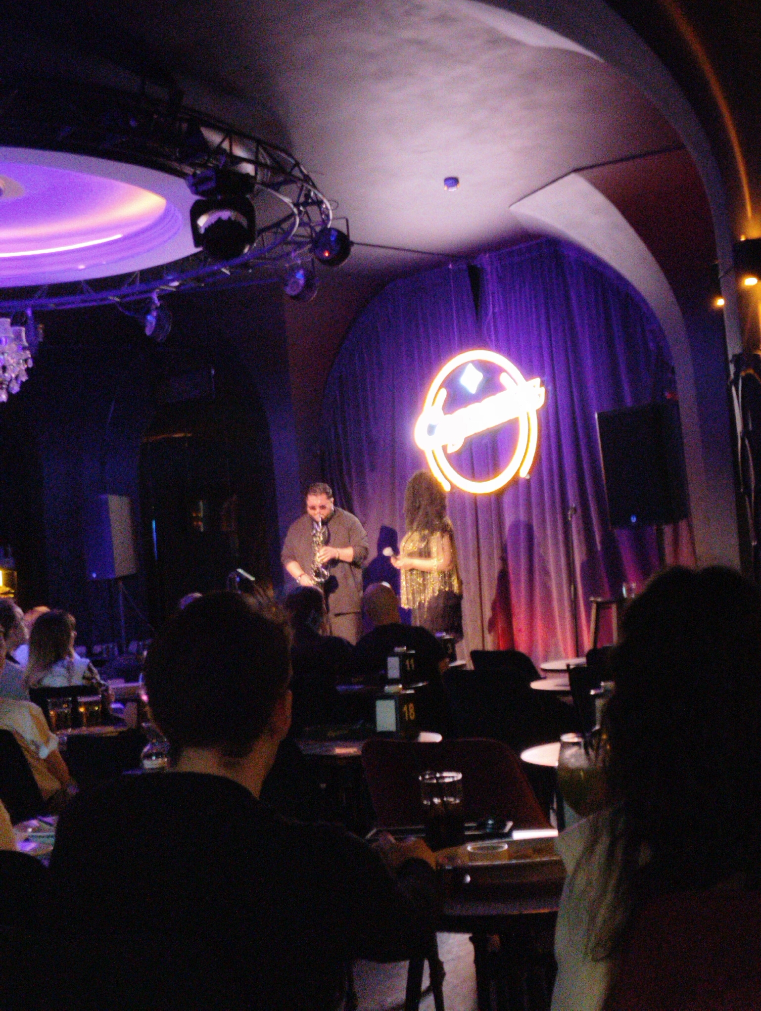 The image shows a jazz club in Moscow, Russia. A man is playing a saxophone on stage, while a woman stands next to him. The stage has a large circular neon sign behind them. The audience is seated at tables, watching the performance. The lighting is dim, with a spotlight on the stage. There are overhead lights with a violet hue. The tables have table numbers visible.