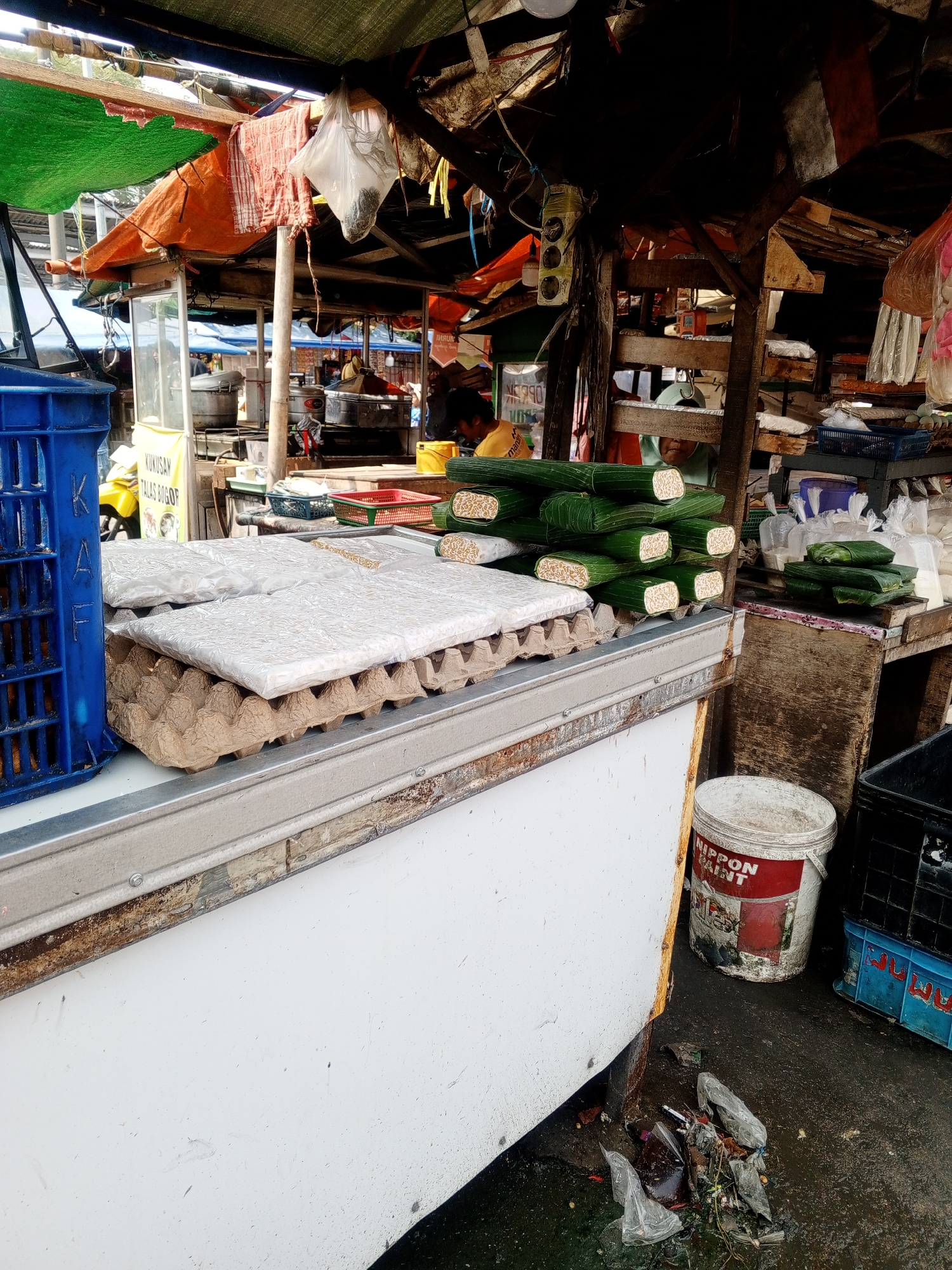 An outdoor market stall in Depok, Indonesia features a display of packaged tempeh. In the foreground, a white refrigerator unit is topped with an egg carton holding several flat, white packages of tempeh. To the right of these packages, a stack of tempeh wrapped in green banana leaves is visible. A blue crate is situated to the left of the tempeh. In the background, the stall's canopy is constructed from corrugated metal and tarpaulin, with various items and food preparation equipment visible. A white bucket with a red label that reads "NIPPON PAINT" is placed on the ground near the stall, with a pile of refuse nearby. The ground is wet and appears to be concrete. The scene depicts a typical street market environment with goods for sale and everyday operational elements.