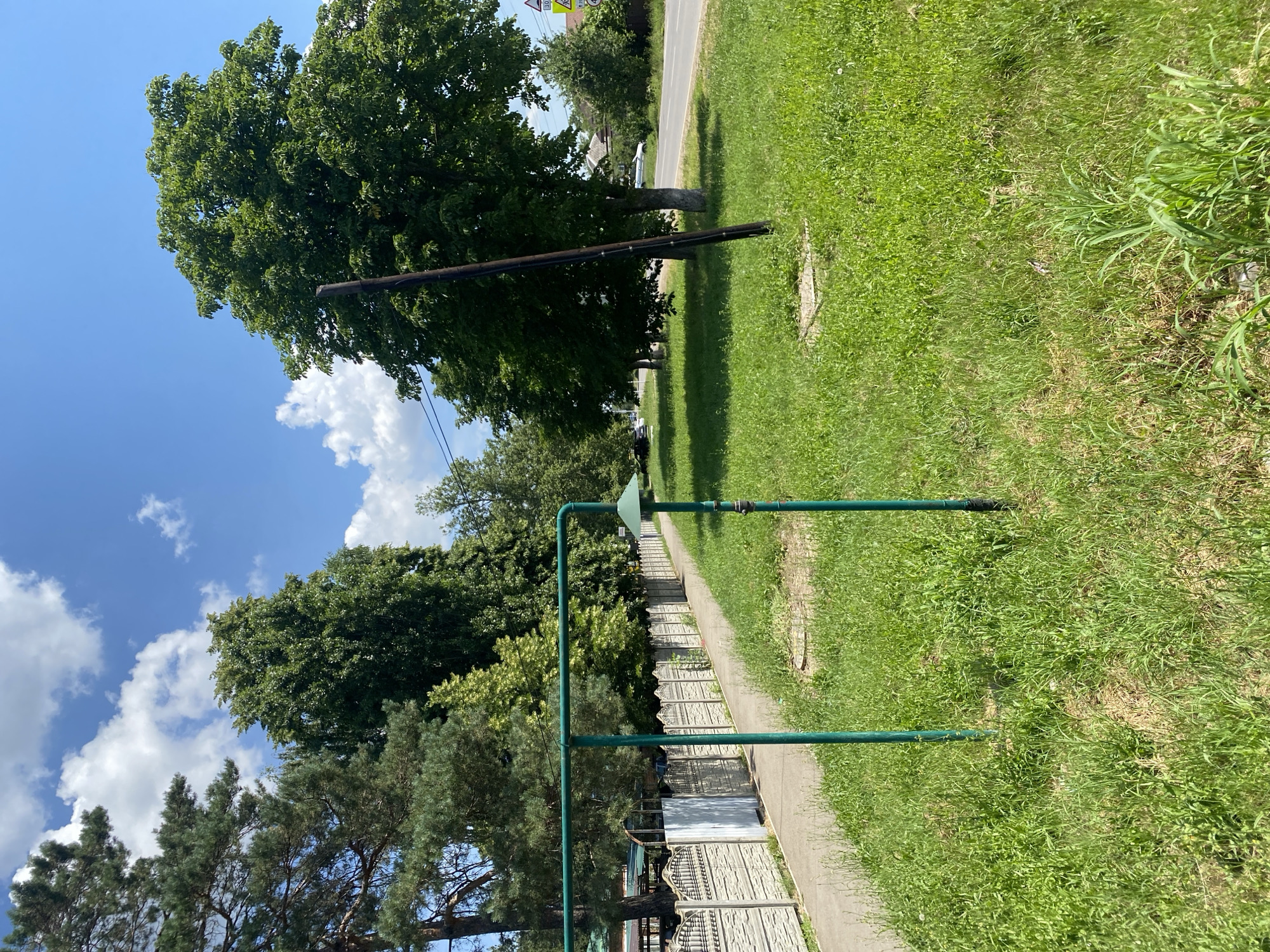 The image shows a bright, sunny day with a clear blue sky dotted with fluffy white clouds. The scene is a grassy area next to a paved pathway. To the left, large, lush green trees dominate the landscape, with one particularly tall tree in the foreground. A thin, dark pole extends upwards from the ground among the trees, with visible wires attached. A green pipe system is also prominent, running horizontally and then upwards, topped with a green conical fitting.
Beyond the pathway, a decorative concrete fence lines a property. In the distance, a road can be seen with some trees and buildings along its edge. The overall setting appears to be a suburban or rural area in Russia. There are no people visible in the image. The grass is green and well-maintained, suggesting it's a pleasant day. The lighting indicates it is daytime, likely late morning or early afternoon. There is no discernible text in the image.