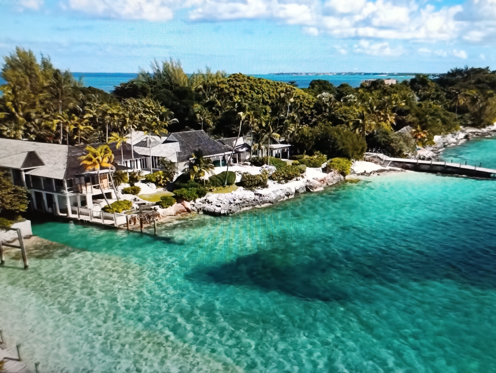 An aerial view captures a luxurious waterfront property situated on a tropical coastline. The scene is dominated by the vibrant turquoise waters of the ocean, which lap against a rocky shore. Several modern, low-lying buildings with grey roofs are nestled amongst lush green palm trees and dense foliage, suggesting a secluded and upscale resort or private estate. A wooden pier extends from the shore into the clear water, and the sandy beach is visible in some areas. The sky above is a bright blue, dotted with fluffy white clouds. There are no people or animals visible in the frame. The image depicts a serene and picturesque tropical setting, possibly a holiday destination. The provided location of Dubăsari town council, Moldova, is inconsistent with the visual evidence of a tropical island environment.