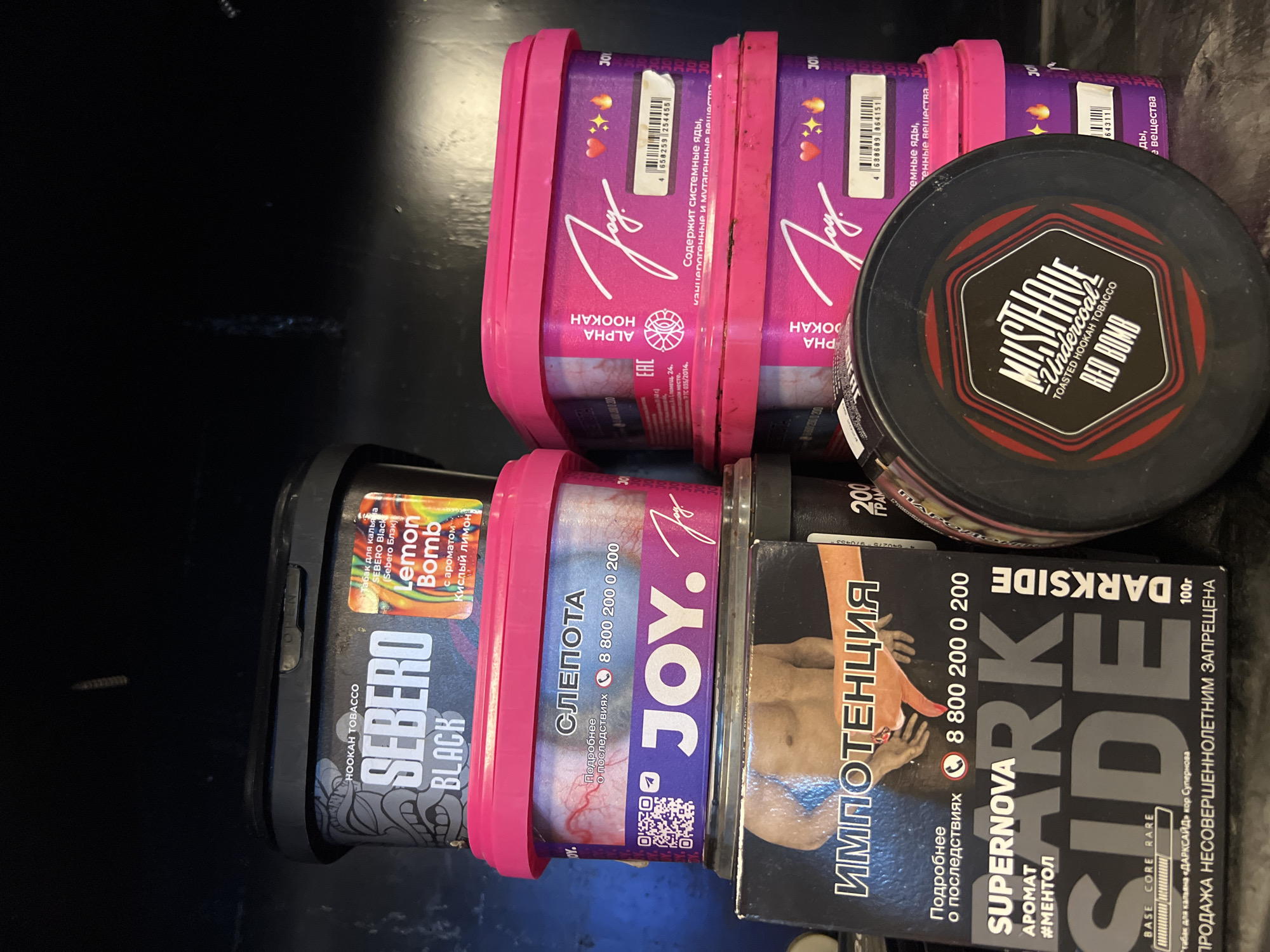 The image shows several containers of hookah tobacco. There are two pink containers labeled "JOY ALPHA HOOKAH", one black container labeled "SEBERO BLACK Lemon Bomb", one black container labeled "DARKSIDE SUPERNOVA" with an image of a bodybuilder, and one black round container labeled "MUSTHAVE Undercoal RED BOMB". They are stacked in a pile. The image does not provide information about a location in Đà Nẵng, Vietnam.