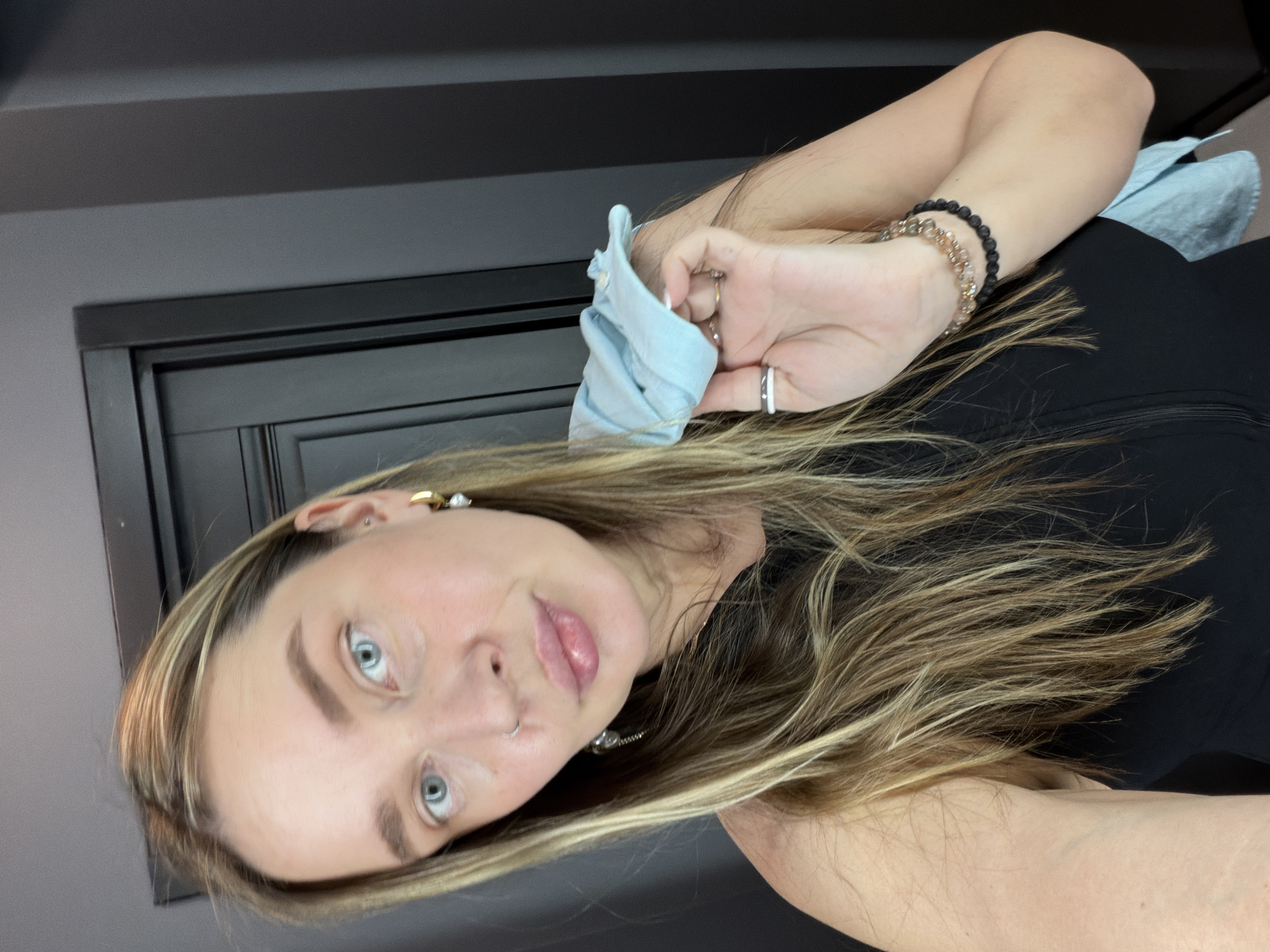 The image shows a woman with blonde hair and blue eyes looking at the camera. She is holding a light blue cloth in her hand. She is wearing a black top and has bracelets on her wrist. The background is a gray wall with a black door frame. There is no indication of the location being Moscow, Russia, or any other specific city.