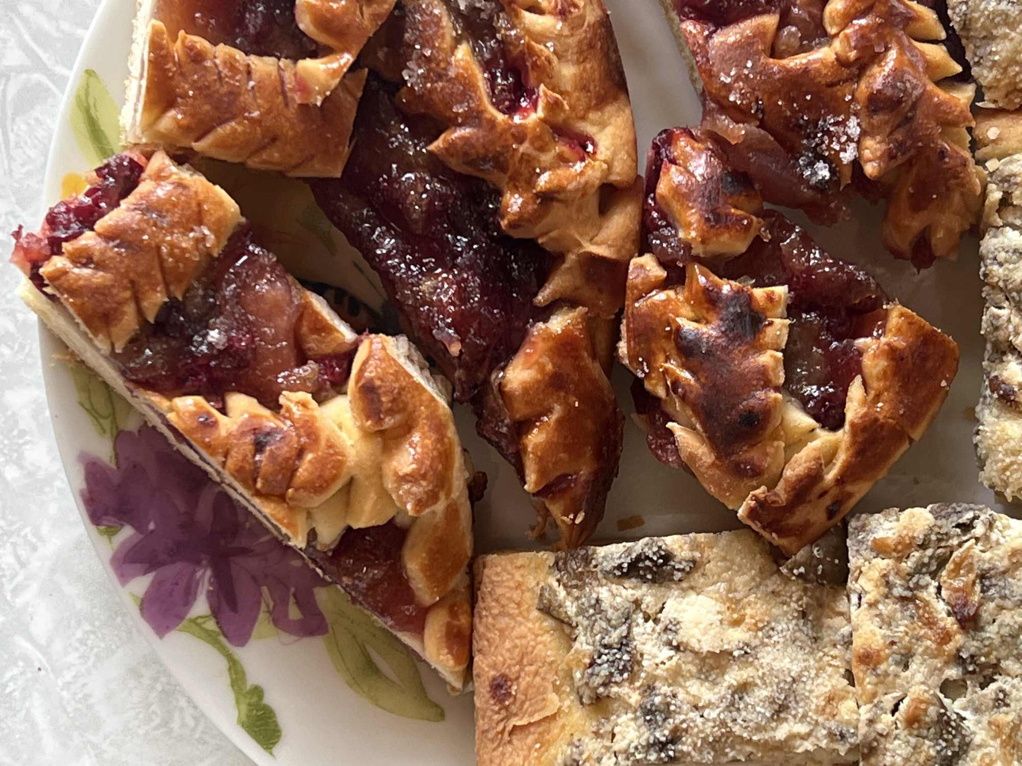 The image shows a plate of baked goods. Some pieces are wedge-shaped, filled with a dark red jam, and topped with decorative pastry. Other pieces are rectangular and covered in a sugared topping with visible raisins. The plate is white with floral decorations, including a purple flower. The background is a neutral, textured surface. The location of Чернышово, Russia is not evident from the image.