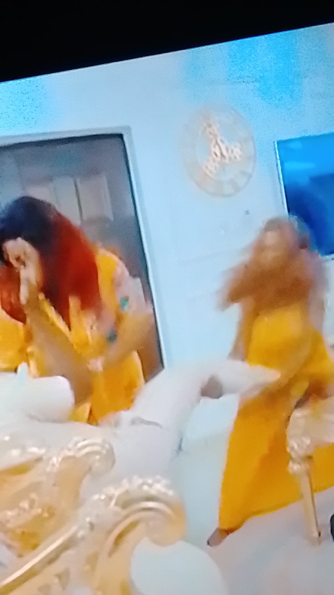 Two women, possibly engaged in a playful pillow fight, are captured in a slightly blurry, overexposed indoor scene. Both women are wearing yellow garments. The woman on the left, with long, dark hair tinged with red, is holding a white object, likely a pillow, and her head is tilted down. The woman on the right, with lighter brown hair, is in motion, also appearing to be holding a pillow. The background suggests a domestic setting, with a wall, a door frame, and a decorative clock on the wall. A ornate, plush sofa or furniture is partially visible in the foreground. The lighting is bright, contributing to the overexposure.