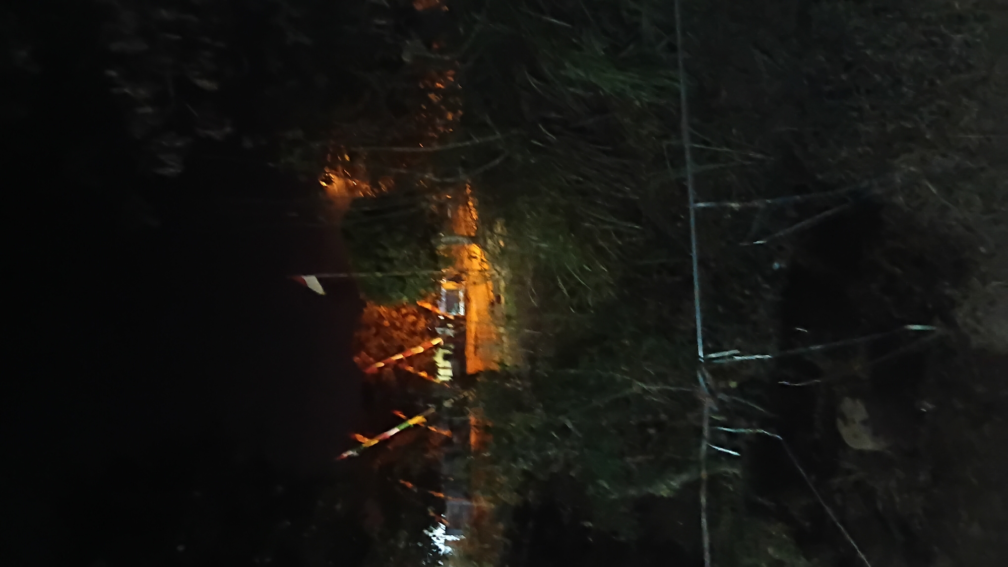 The image depicts a night scene in Prabumulih, Indonesia, featuring an Indonesian flag flying on a pole. In the background, several structures with pointed roofs are illuminated by artificial lights, suggesting they might be traditional houses or a cultural landmark. The foreground is dominated by dark, dense vegetation, with a metal fence running horizontally across the bottom of the frame. The overall atmosphere is dimly lit, with the colors muted by the darkness.