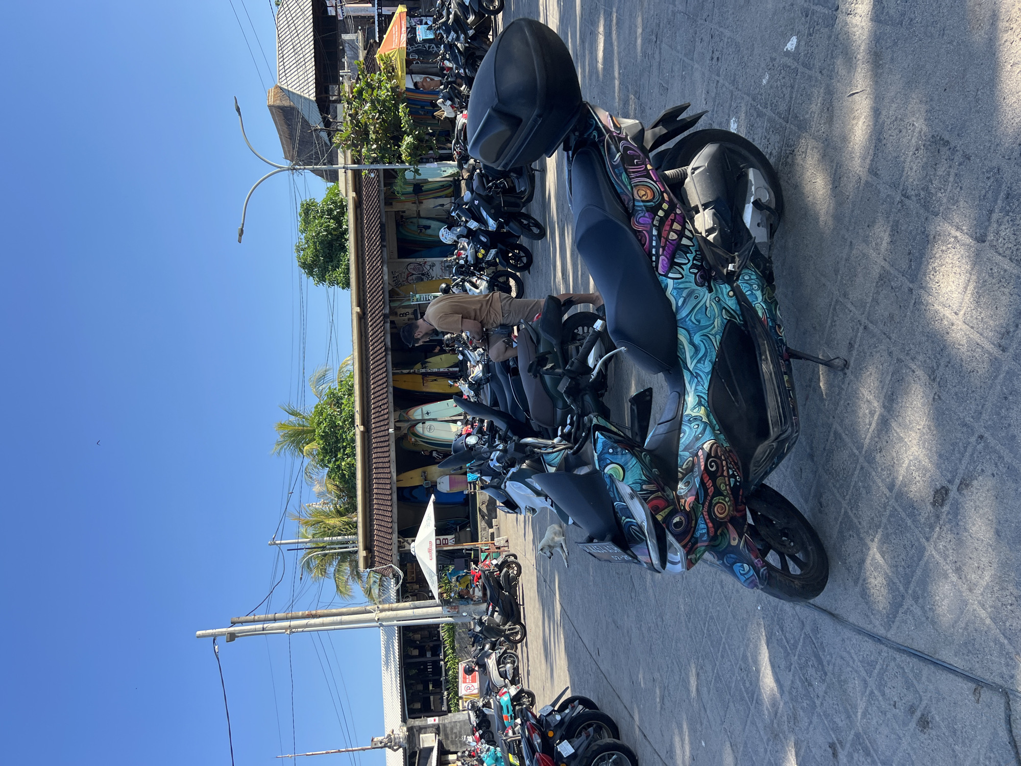 A parking lot in the city of North Kuta, Indonesia, is filled with parked scooters. In the foreground, a scooter with a vibrant, colorful mural design is prominently displayed, with its front wheel and part of its body visible. Behind it, many other scooters are parked in rows, some with riders, and a white dog is lying on the ground. In the background, a building with a thatched roof is visible, with surfboards leaning against it. Power lines and street lights are also present in the scene, indicating an urban or semi-urban environment. The sky is clear and blue, suggesting a sunny day.