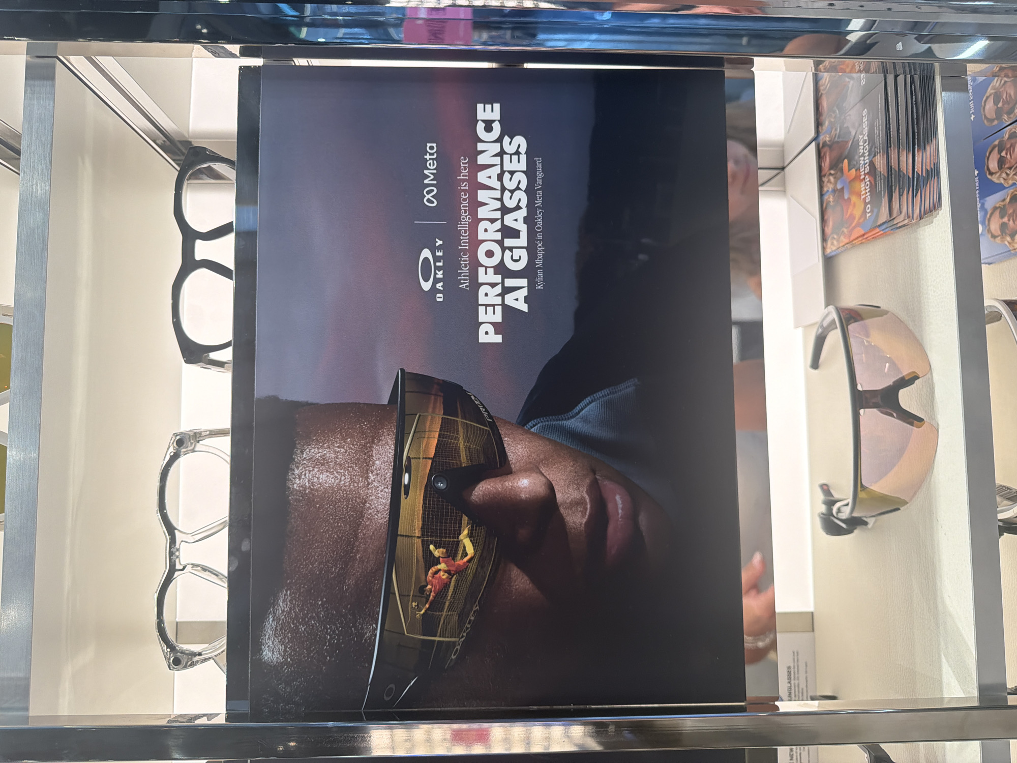 This is a medium shot of a retail display of eyewear, likely in a store in Greater London, United Kingdom. The display features a large advertisement poster for "Oakley Meta Performance AI Glasses," showing a close-up of a person's face wearing the glasses, with a reflection of a soccer player in the lenses. The poster also includes the Oakley and Meta logos, and text that reads "Athletic Intelligence is here."
Below the poster, on a shelf, are several pairs of sunglasses and a stack of brochures. The brochures have an image of two women wearing sunglasses and the text "THE NEW WAY TO SHOP SUNGLASSES." In the background, on a higher shelf, there are two pairs of eyeglasses. The overall environment appears to be a well-lit retail space with a modern aesthetic, featuring metal shelving.