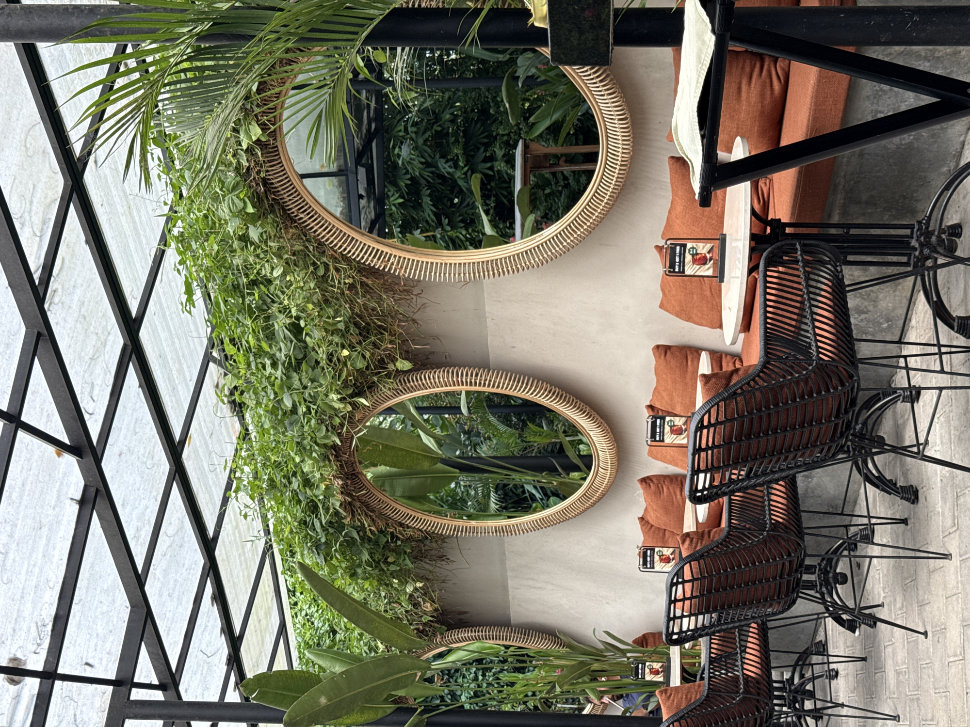 The image captures an outdoor seating area, likely a cafe or restaurant, located in Ubud District, Indonesia. The scene showcases a row of rattan-framed, oval-shaped mirrors adorned with abundant greenery, creating a natural and inviting aesthetic. Adjacent to the mirrored wall, there is a seating arrangement with comfortable, terracotta-colored cushions and black woven chairs arranged around small tables. The presence of menus or promotional cards on the tables indicates a dining establishment. Above, a partial view of a black metal frame structure suggests a covered or semi-covered area, offering shade and protection from the elements. The overall ambiance is relaxed and harmonious, blending natural elements with modern design, characteristic of Ubud's distinctive style.