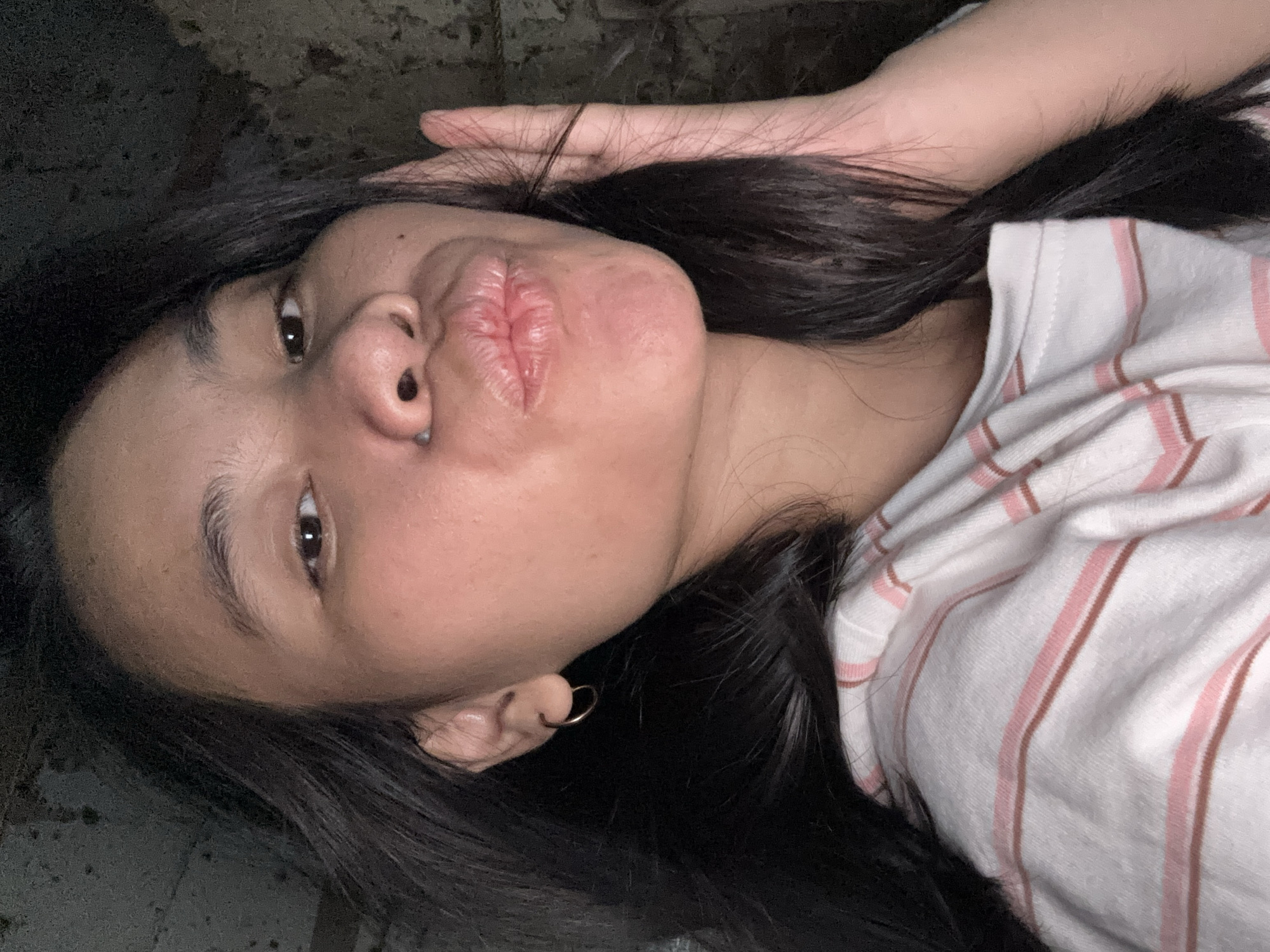 The image is a close-up, front-facing selfie of a young woman. She has dark hair that frames her face and is wearing a white t-shirt with thin pink and brown stripes. Her left hand is raised to her face, with her fingers touching her cheek and hair. Her lips are pursed, as if for a kiss. The background is dark and appears to be a textured wall. The lighting is dim, suggesting an indoor setting at night. There is no visible indication of the location within East Jakarta, Indonesia.