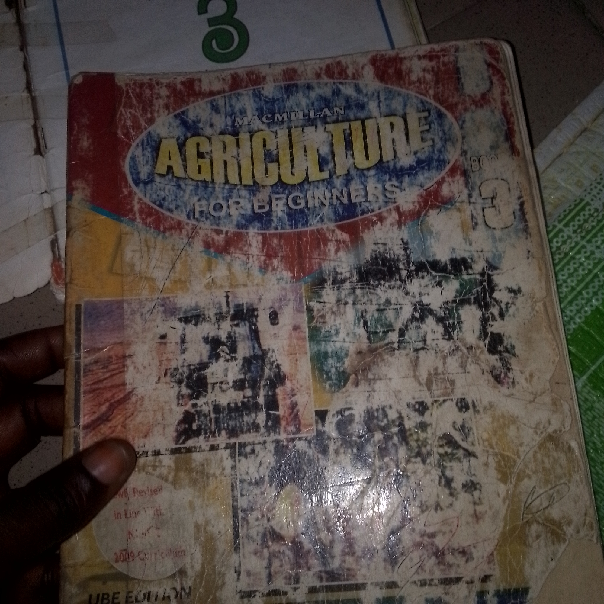 The image shows a close-up, eye-level, indoor shot of a worn and damaged book. The book appears to be titled "AGRICULTURE FOR BEGINNERS" with "MACMILLAN" printed above it. The cover is heavily creased, torn, and stained, with sections of the original design obscured by grime and wear. A hand with dark skin is visible on the left side of the frame, obscuring a portion of the book. Part of the book's spine is visible on the right, showing a green patterned material underneath, possibly a table or shelf. Faint imagery and text are discernible on the book cover, including what looks like a circular sticker with text in red and black. The book itself appears to be lying on a surface, with the green patterned material and possibly another surface or object visible to the right and behind. The lighting suggests an indoor setting. There is no indication of the city of Jalingo, Nigeria, in the image.