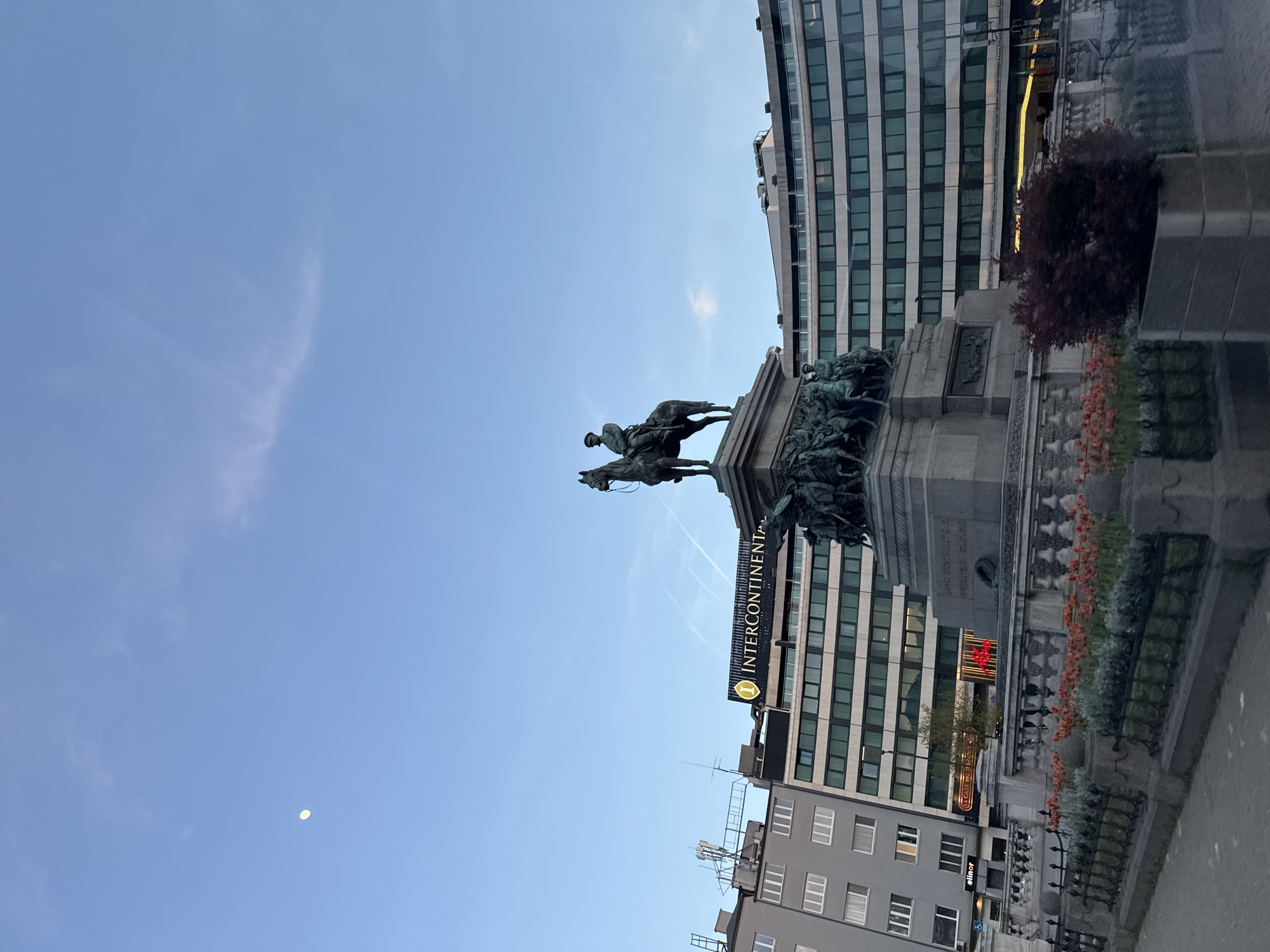 An outdoor, wide-angle shot captures a prominent equestrian statue against a backdrop of modern buildings and a clear sky in Sofia, Bulgaria.
The central subject is a large bronze equestrian statue depicting a historical figure, likely a military leader, mounted on a horse. The statue stands atop a grand stone pedestal, which is intricately carved with relief sculptures of multiple figures in dynamic poses, suggesting historical or allegorical scenes.
In the background, several multi-story buildings rise into the sky. A large, light-colored building with many windows stands directly behind the monument. To its left, a darker building prominently features an illuminated sign displaying the word "INTERCONTINENTAL" in capital letters, accompanied by its distinctive logo. Below this, on a lower building, the text "CAFE DE L'OPERA" is faintly visible. Further to the left and below, another sign reads "elinor".
The sky is a bright, clear blue with a few wispy clouds. A thin, crescent moon is distinctly visible in the upper left quadrant of the frame, indicating either early morning or late afternoon/early evening. The lighting suggests a soft, ambient daylight.
At the base of the statue's pedestal, there's an ornate railing or wall lined with planters. These planters hold vibrant reddish-orange flowers and green foliage, adding a touch of color to the urban environment. A dark red leafy tree is also visible to the right of the monument. The scene depicts a significant public monument within an urban landscape.