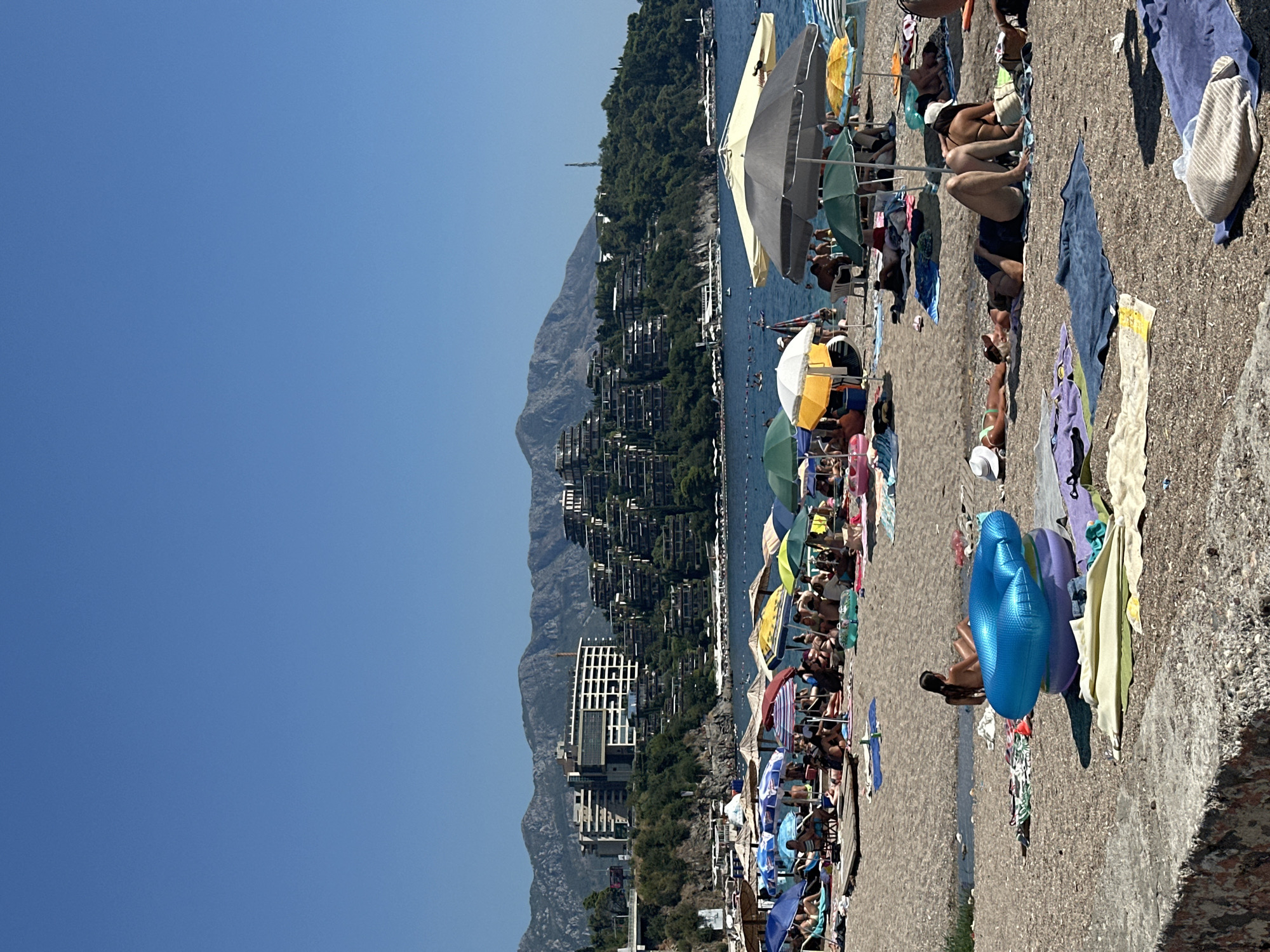 This image captures a crowded beach in Budva, Montenegro, likely during the summer season, under a clear blue sky. The foreground features a rough stone wall, followed by a beach covered in sand and pebbles. A multitude of beach umbrellas in various colors – yellow, blue, green, and patterned – are spread across the sand, providing shade for numerous sunbathers. People are seen lying on towels and mats, some facing the sun, others reading or chatting.
In the middle ground, the calm blue sea stretches towards the horizon. Several boats are visible on the water, and a few people are swimming. Behind the beach, a lush green hillside is dotted with trees and a distinctive modern resort complex. The buildings are designed in a terraced, stepped architectural style, creating a unique visual impact against the natural landscape. The resort's white facades and geometric shapes contrast with the surrounding greenery and the rugged, grey mountains that form the backdrop of the scene. The overall impression is one of a popular tourist destination, bustling with activity and enjoying a sunny day.