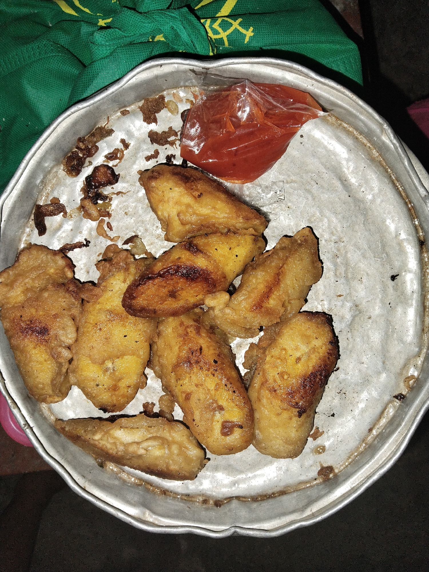 A metal plate holds fried plantains and a small plastic bag of reddish sauce. The setting appears to be a simple, informal environment. The exact location is Unknown, Unknown, as no location information is available.
