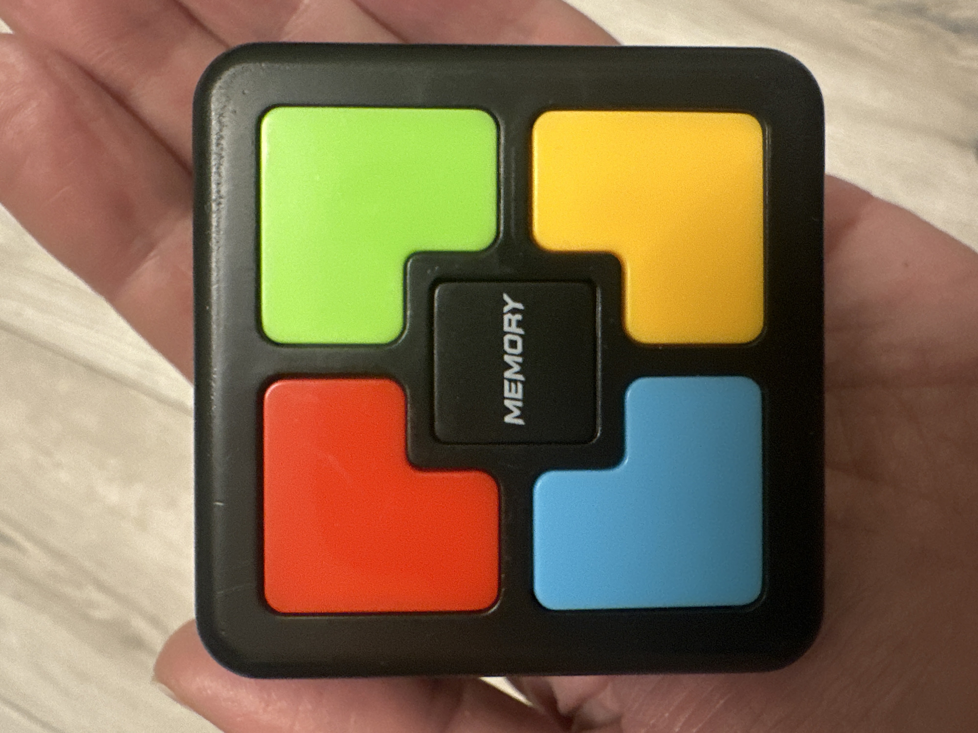 A close-up photograph shows a handheld electronic memory game. The device is square-shaped with a black casing. It features four colored buttons (green, yellow, red, and blue) arranged around a central black button labeled "MEMORY". The game is held in someone's hand. The background is a blurred, neutral color, possibly a wooden surface. The image contains no identifying landmarks, people, or indications of a location within the city of Riga, Latvia, or any other specific locale.