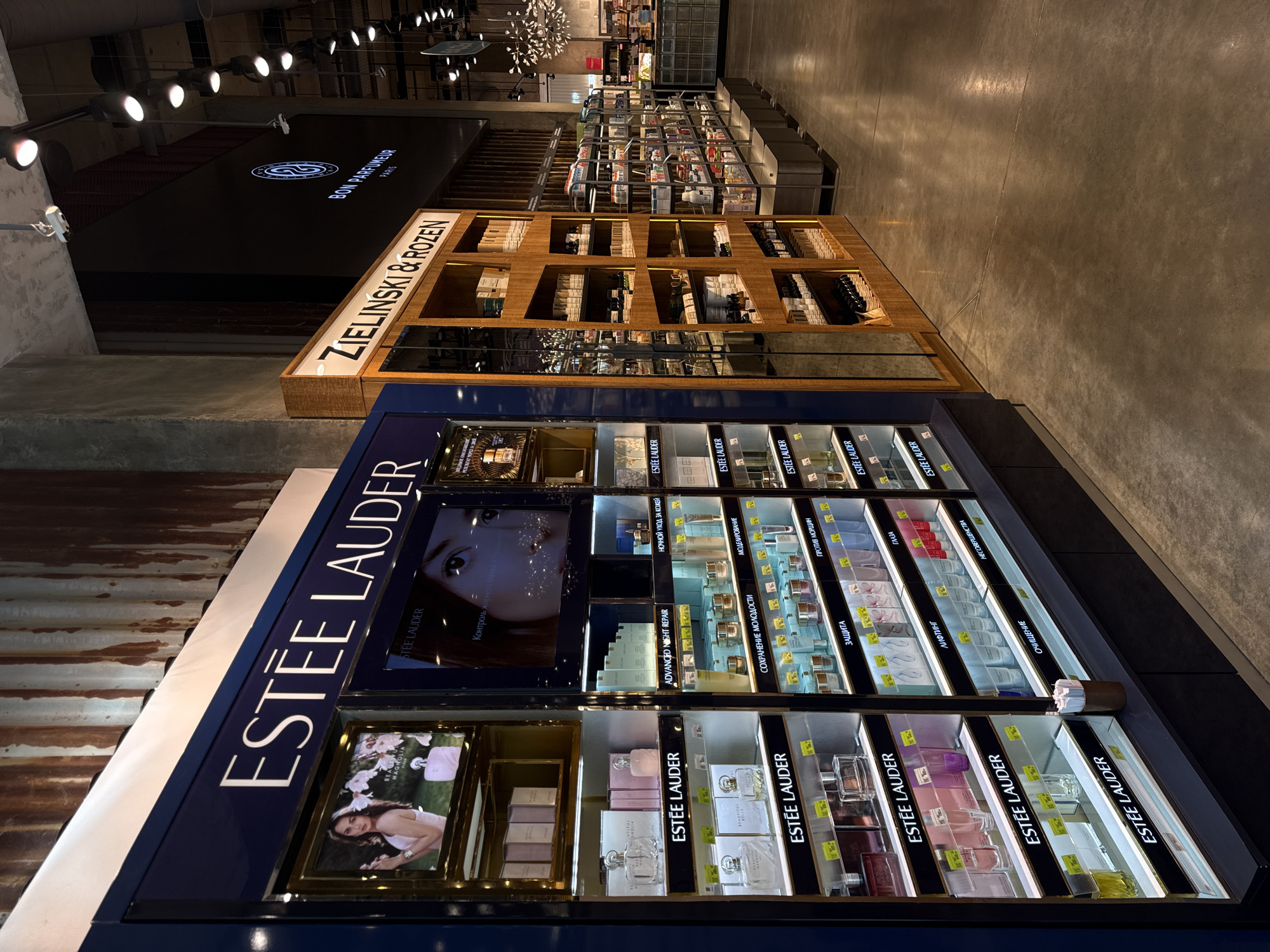 The image displays the interior of a modern beauty and fragrance store section in Perm, Russia. The scene is brightly lit by numerous overhead spotlights, illuminating various retail displays. The flooring is a neutral, polished concrete, and the store exhibits a contemporary design with visible structural elements and high ceilings.
In the foreground and to the left, a large, dark blue display cabinet prominently features the "ESTÉE LAUDER" brand in white, uppercase letters. This display showcases a range of skincare and perfume products. Visible product lines and categories include "ADVANCED NIGHT REPAIR," "BEAUTIFUL," and sections labeled with Russian text: "НОЧНОЙ УХОД ЗА КОЖЕЙ" (Night skin care), "СОХРАНЕНИЕ МОЛОДОСТИ" (Youth preservation), "МОДЕЛИРОВАНИЕ" (Modeling/Sculpting), "ЗАЩИТА" (Protection), "ПРОТИВ МОРЩИН" (Anti-wrinkle), "ЛИФТИНГ" (Lifting), "ГЛАЗА" (Eyes), "ОЧИЩЕНИЕ" (Cleansing), and "НЕСОВЕРШЕНСТВА" (Imperfections). A screen on the display also mentions "Контроль жирного блеска" (Oil control).
Further back and to the right, a striking wooden display unit with multiple compartments and glass shelves features the brand "ZIELINSKI & ROZEN." This unit holds numerous bottled products, likely fragrances or personal care items. Beyond this, additional shelves and displays with various products are visible, indicating a comprehensive beauty retail space.
Above, an illuminated sign on a dark surface reads "BON PARFUMEUR PARIS," suggesting another brand or dedicated section. The environment is clean and organized, designed for browsing and purchasing beauty products. No customers or staff are visible in the frame. The time of day is not discernible from the indoor lighting.