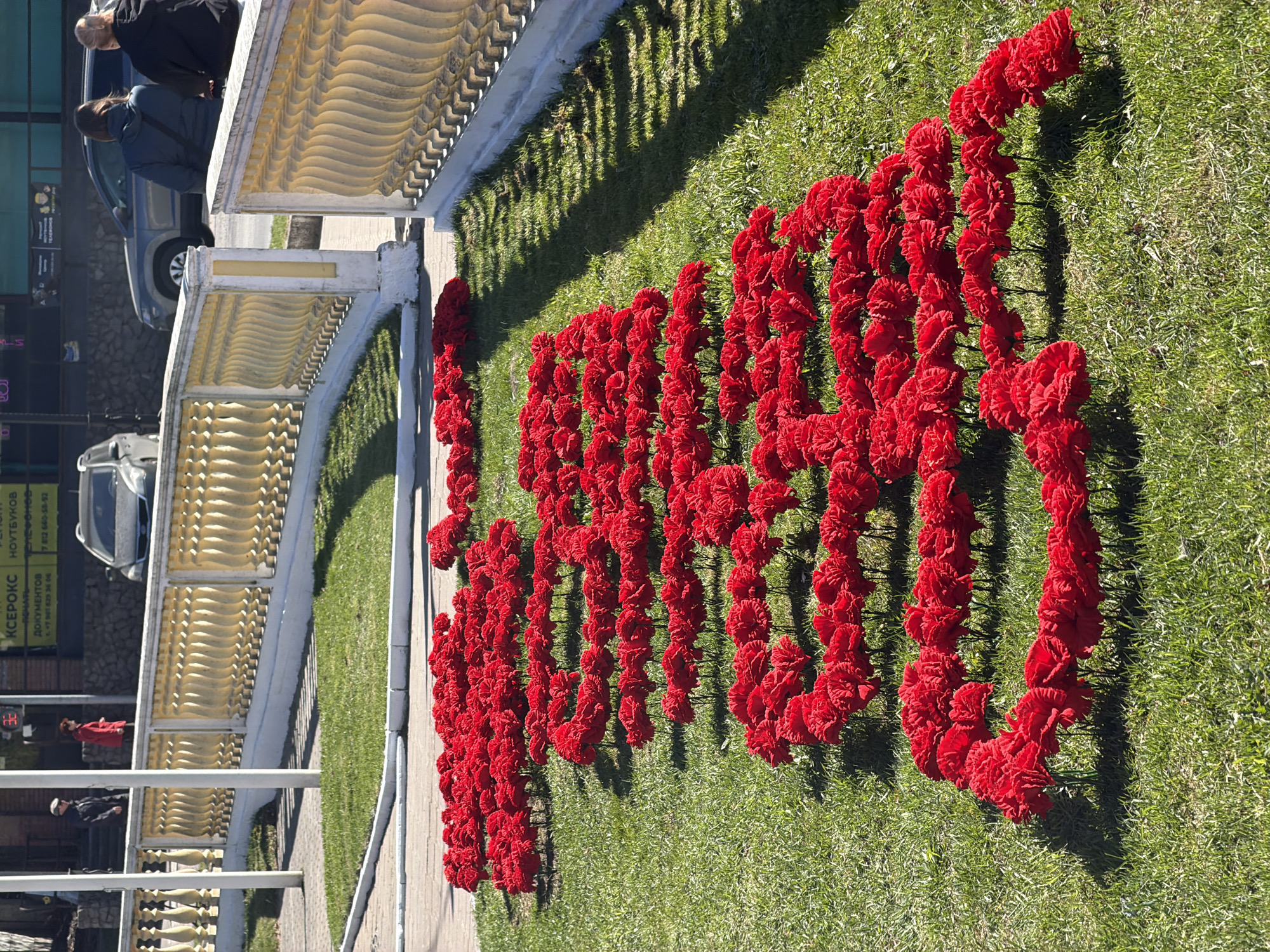 The image shows an arrangement of red flowers, likely carnations, forming the Cyrillic letters "СПБ," which is the abbreviation for Saint Petersburg, Russia, on a green lawn. A light-colored, ornate railing or balustrade runs along the edge of the lawn, casting shadows onto the grass. In the background, buildings and vehicles are visible. There are people walking along the sidewalk next to the balustrade. A sign with Cyrillic script is visible on one of the buildings. The flowers seem to be deliberately placed to create this memorial or commemorative display on the lawn.
