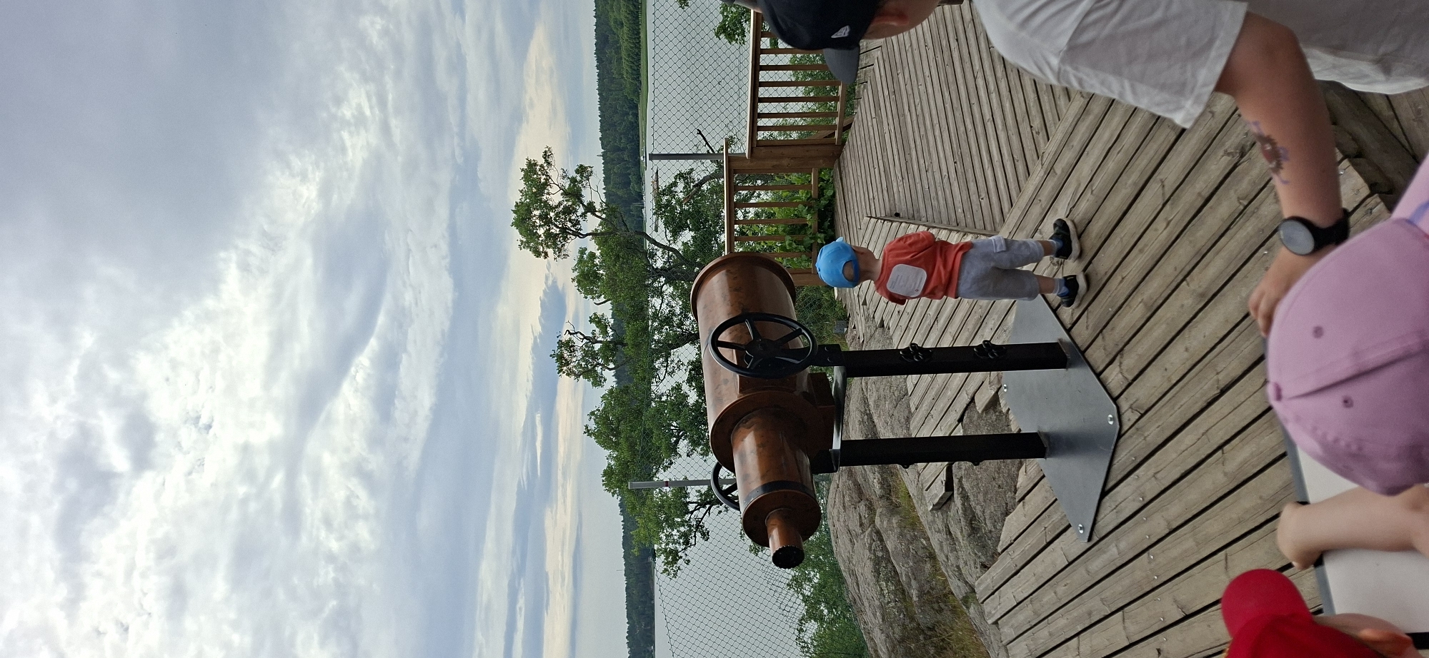 The image depicts a view from a wooden platform overlooking a body of water, likely the archipelago surrounding Naantali, Finland, on a cloudy day. In the foreground, a large, copper-colored telescope-like object is mounted on a black stand. A young child wearing a blue hat and an orange t-shirt stands facing away from the camera, looking towards the water. To the right, the back of another person's head is visible, wearing a black cap. In the lower portion of the image, the backs of two other children's heads are visible, one wearing a red cap and the other a pink cap. The platform is made of wooden planks and is elevated, suggesting it is a viewpoint. Trees and a chain-link fence line the edge of the platform overlooking the water. The distant shore is lined with trees.