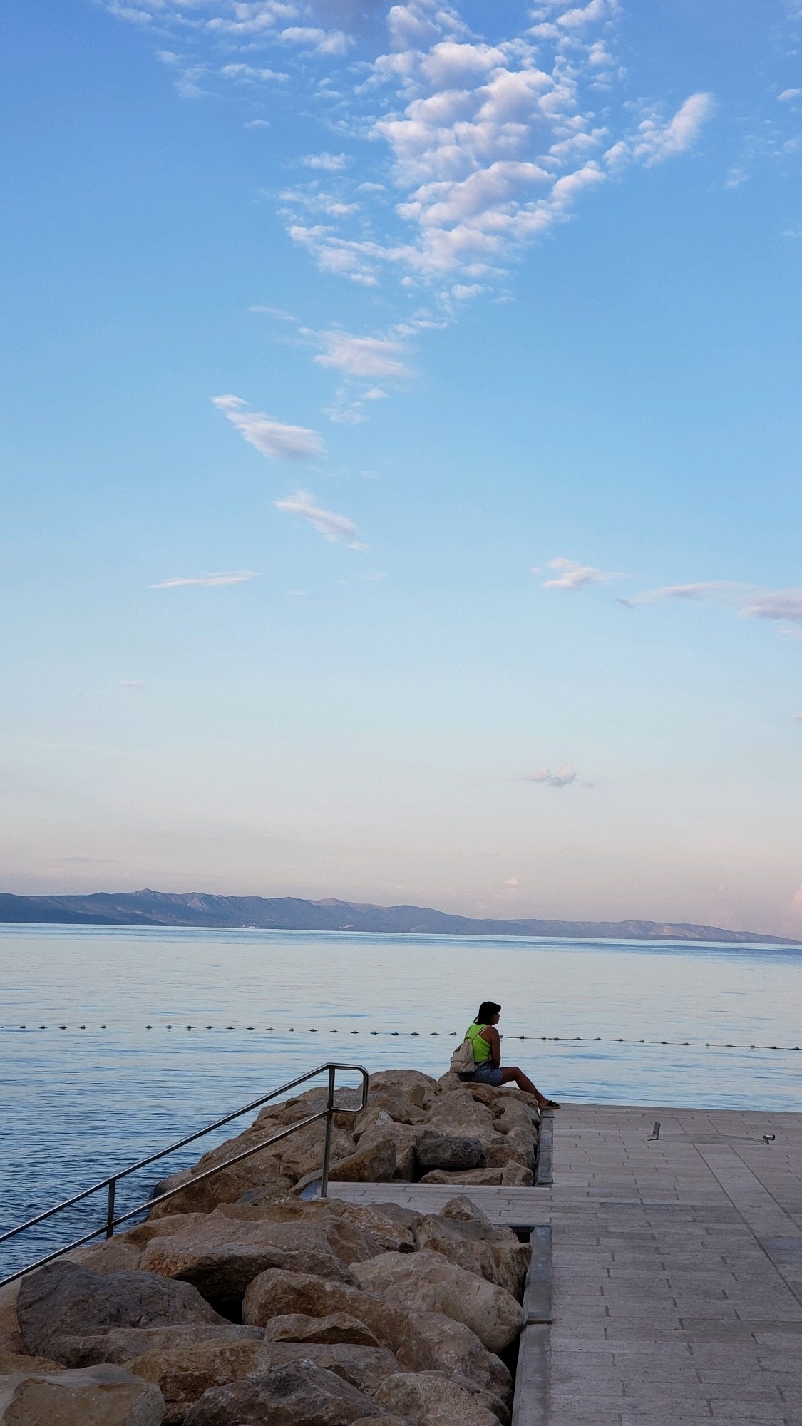 A young woman with dark hair, wearing a neon green tank top and denim shorts, sits on large rocks by the sea in Općina Tučepi, Croatia. She is facing away from the camera, looking out at the water. A light-colored backpack is on her back. The sea is calm, with a line of buoys marking a boundary in the water. In the distance, mountains are visible on the horizon. The sky is blue with scattered white clouds. A paved walkway is to the right of the rocks, and a metal railing leads down to the water. The scene appears to be a tranquil setting at the coast.