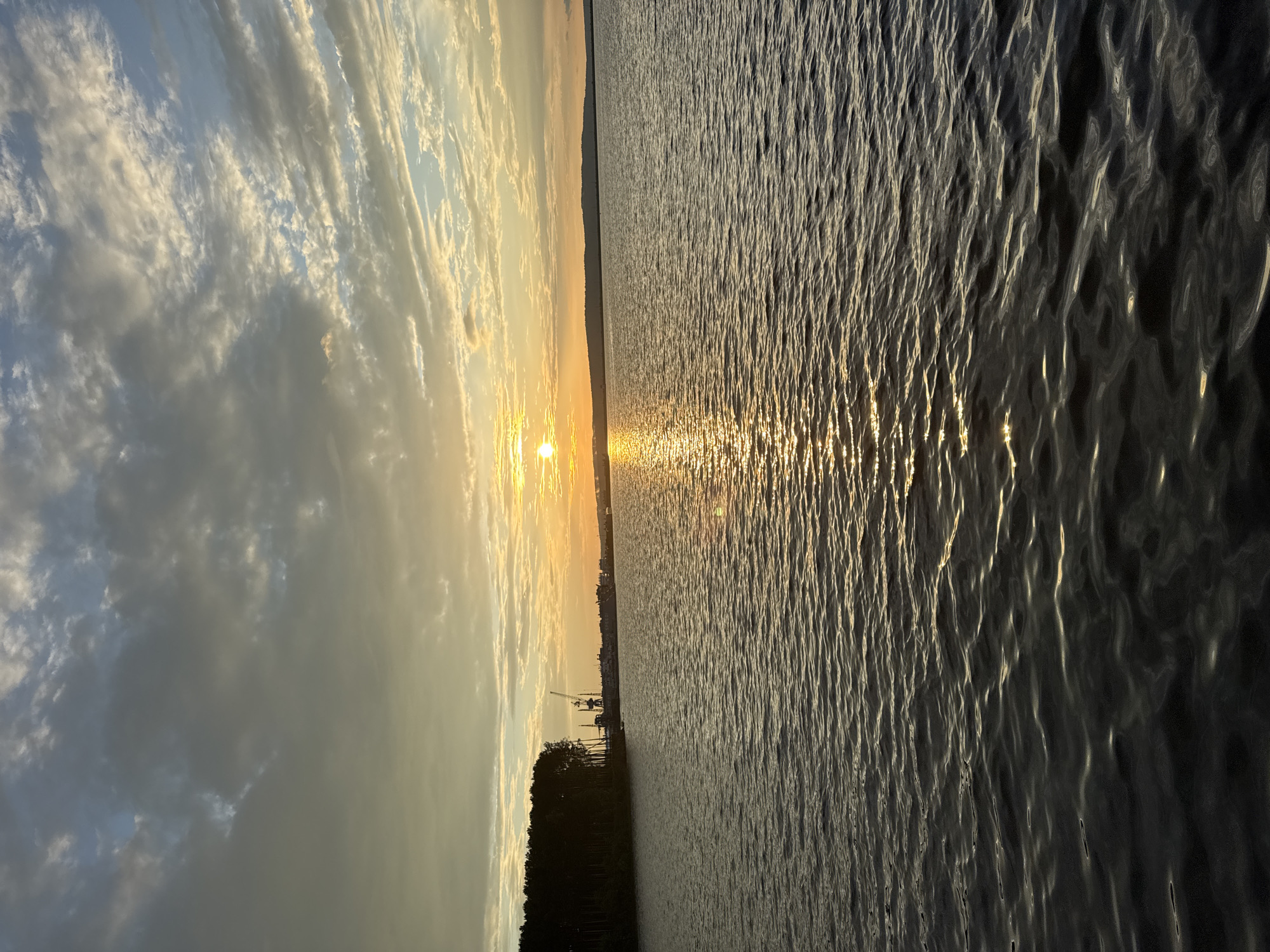 The image depicts a sunset over the Kama River in Chaikovsky, Russia. The sun, a bright orb with a shimmering reflection on the water, is setting near the horizon, casting a warm golden glow across the sky. The sky is filled with a mix of clouds, ranging from dark, heavy masses to wispy, illuminated streaks.
On the left bank of the river, a dense line of dark green trees forms a distinct silhouette against the brighter sky. Beyond the trees and to the right, industrial elements are visible, including cranes and what appear to be ships or port structures. A distant, low-lying landmass is visible on the far horizon. The water in the foreground is rippled, with sunlight reflecting off the crests of the waves, creating a dynamic texture. The overall atmosphere is serene and natural, with a hint of industrial presence on the shoreline. There are no people or animals visible in the scene.