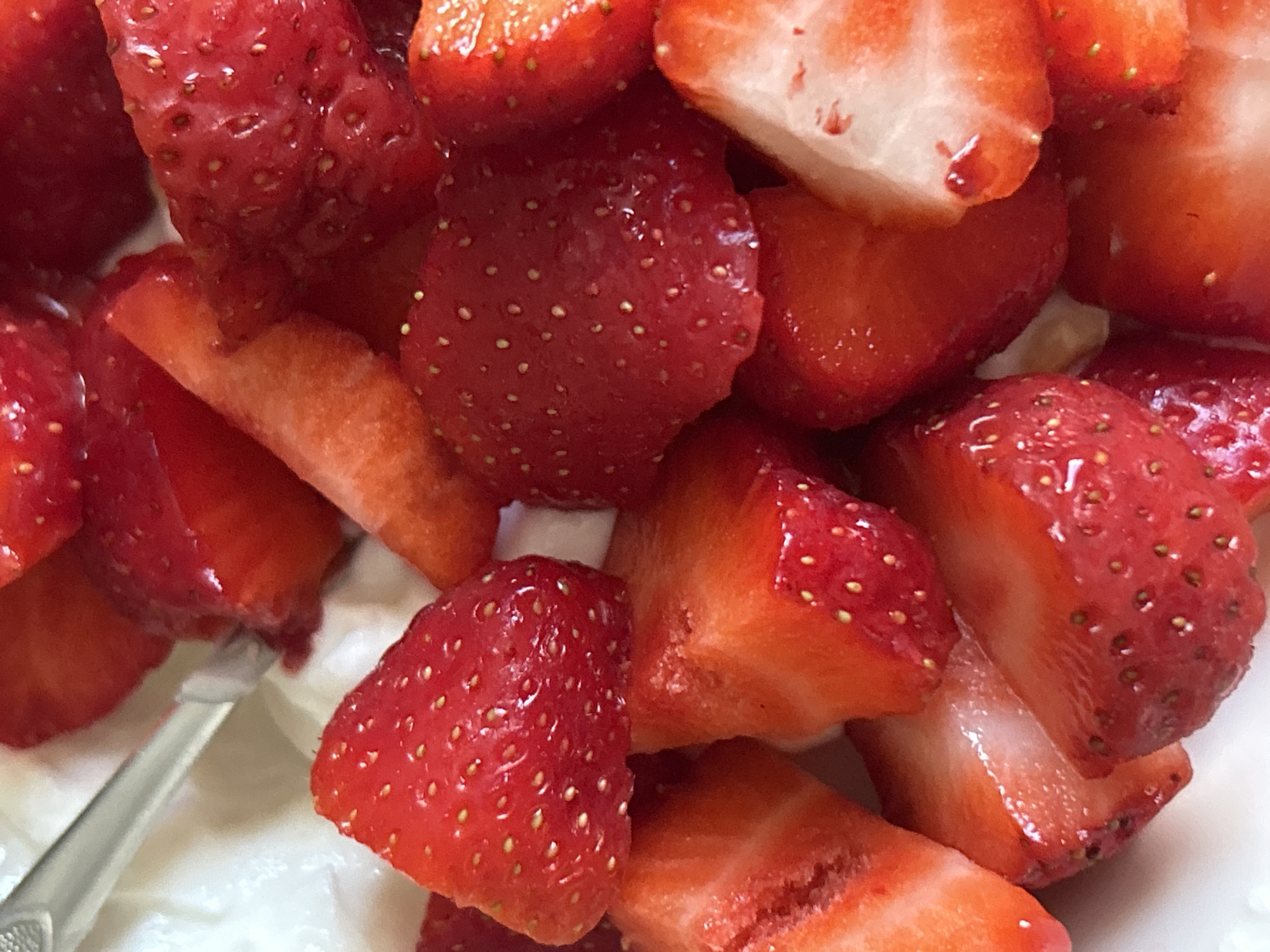 The image shows a close-up view of a bowl filled with sliced strawberries and a creamy white substance, likely yogurt or whipped cream. A silver spoon is partially submerged in the mixture. The strawberries are vibrant red, with some showing their characteristic seeds. The lighting suggests an indoor setting. There is no discernible location within Plovdiv, Bulgaria, or any other city mentioned.