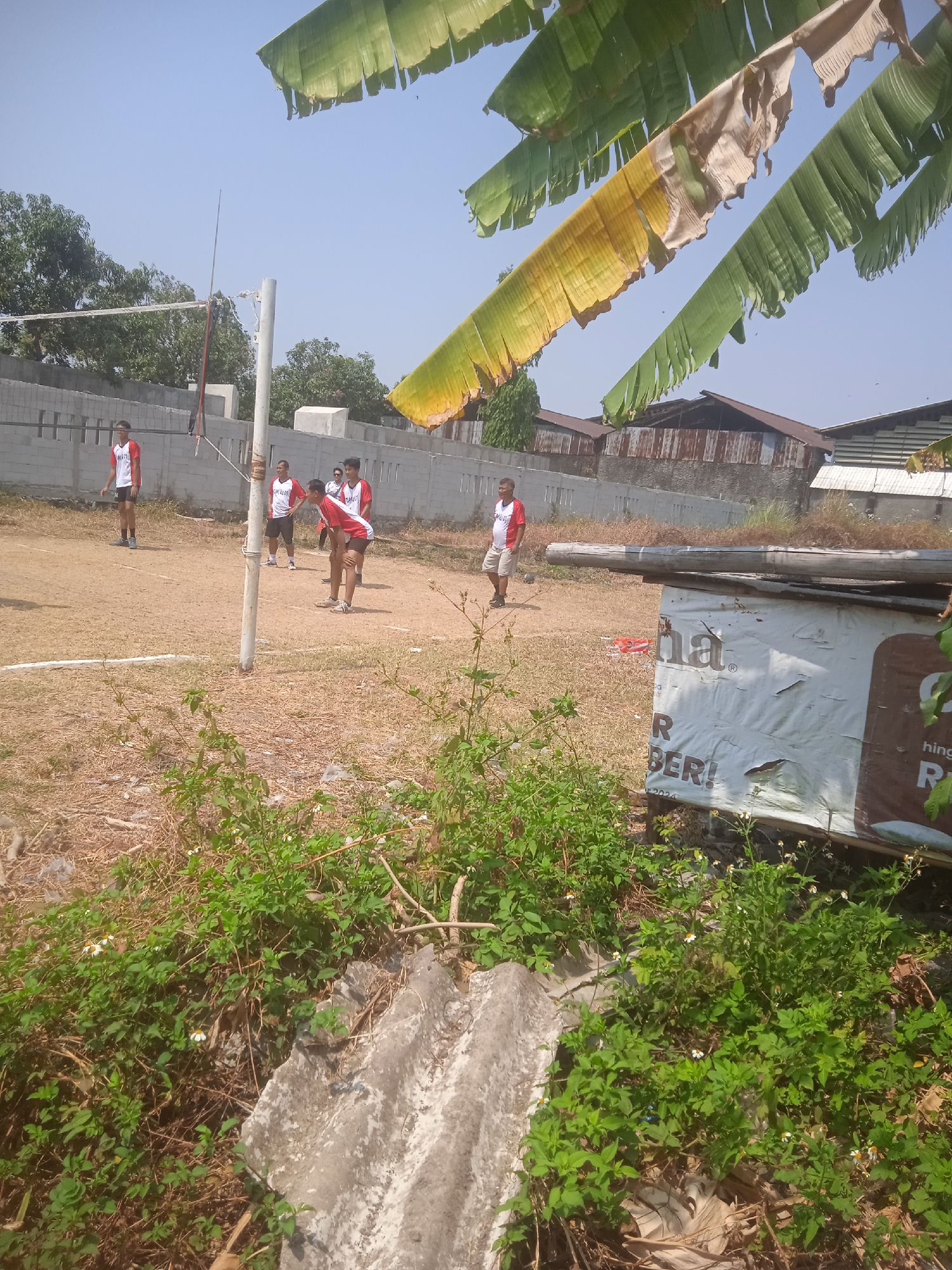 The image depicts an outdoor volleyball court located in Klodran, Indonesia. Several individuals, likely athletes wearing red and white uniforms, are scattered across the dusty court. A volleyball net is visible, stretched between two poles. In the foreground, lush green vegetation and some concrete debris are present, partially obscuring a weathered advertisement billboard. The background shows a line of buildings with corrugated metal roofs, and a clear blue sky overhead. The overall scene suggests a casual sports activity taking place in a semi-urban or rural setting.