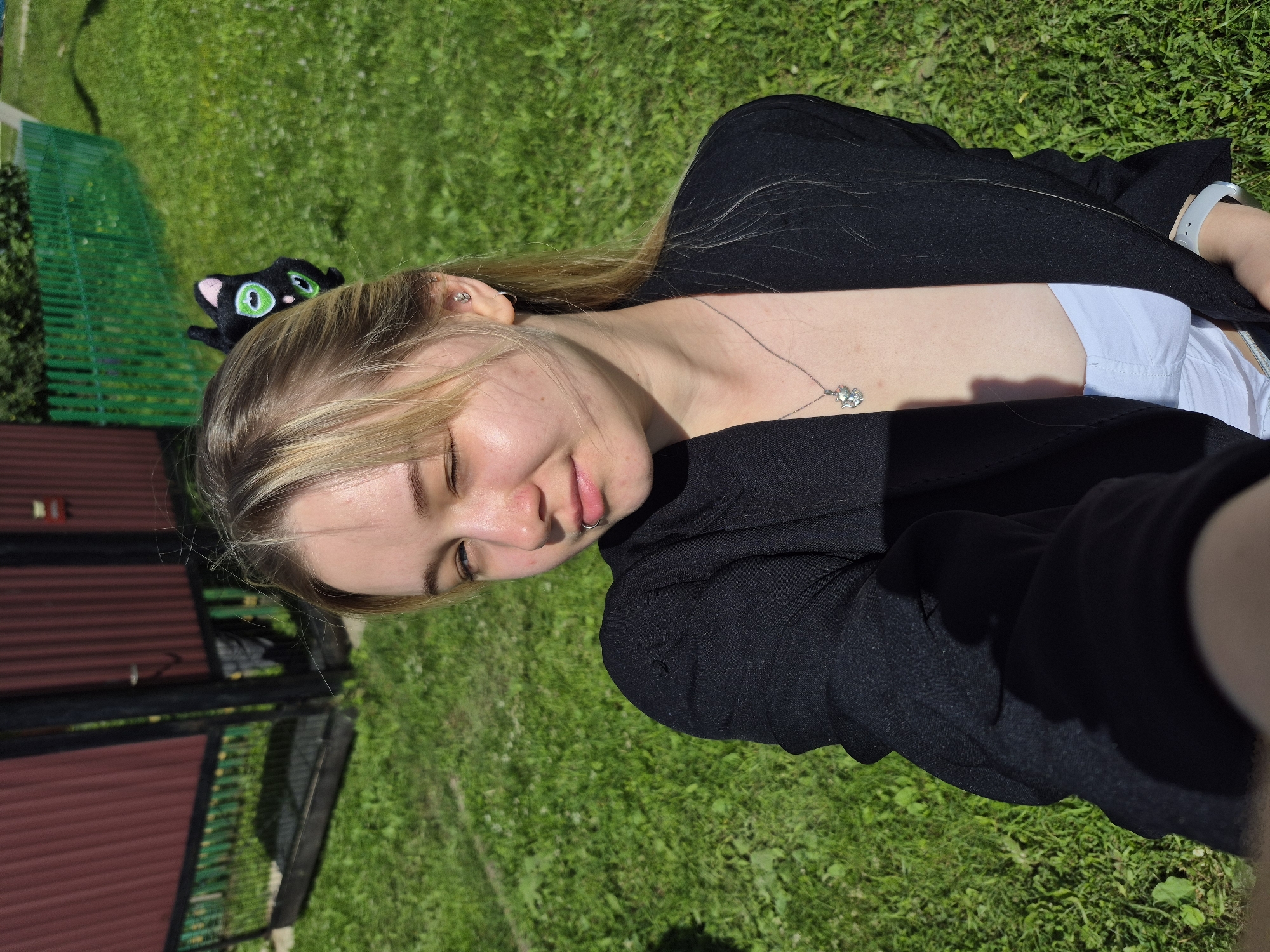 A young woman is captured in a selfie outdoors. She is wearing a black blazer over a white top and a silver necklace with a pendant. Her hair is blonde and pulled back, with a black hair clip shaped like a cat. She has a lip ring and a piercing in her ear. The background shows a green lawn, a red fence, and a glimpse of a building. The lighting suggests it is a sunny day. The image appears to be taken in the city of Sars, Russia, as indicated by the presence of a traditional wooden fence and a corrugated metal structure.