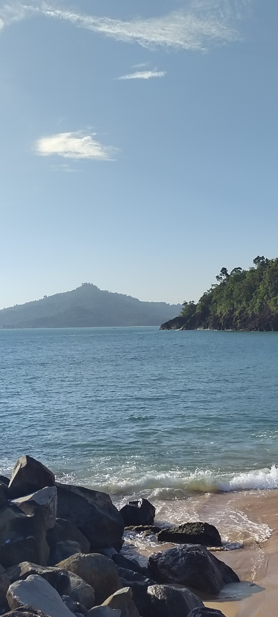 The image depicts a serene coastal scene in the city of Calang, Indonesia. The foreground is dominated by a cluster of large, dark rocks scattered along a sandy beach. Gentle waves are lapping at the shore, creating a foamy white edge where the water meets the sand.
Beyond the immediate shore, a vast expanse of calm, blue ocean stretches towards the horizon. In the middle ground, two islands or peninsulas are visible. The one on the left is a large, tree-covered mountain shrouded in a light haze, appearing distant and majestic. The island on the right is closer, its steep slopes covered in lush green vegetation, with rocky outcrops along its coastline.
The sky above is a clear, pale blue, with a few wispy white clouds scattered across it. The sunlight suggests it is daytime, likely mid-morning or late afternoon, given the soft lighting. There are no people or visible man-made structures in the frame, emphasizing the natural beauty of the location. The overall atmosphere is one of tranquility and natural splendor.