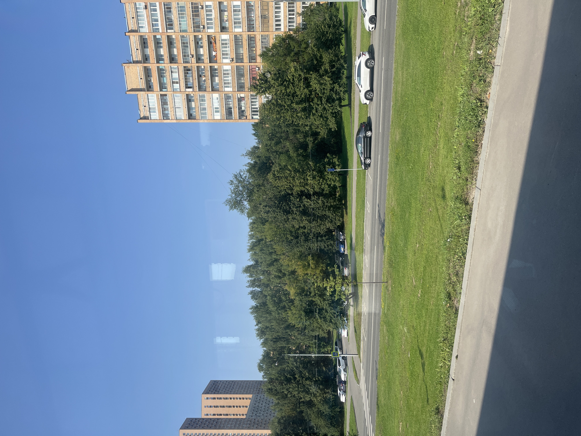 The image depicts a view from a window overlooking a street scene in Moscow, Russia, likely in a residential area. In the foreground, a paved surface, possibly a sidewalk or part of a road, is visible with a distinct shadow cast across it. Beyond this, a strip of green grass with some scattered weeds runs alongside a road. Several cars are parked on the grass area to the left and are also driving on the road. A pedestrian crossing is marked on the road. A line of lush green trees forms a dense barrier behind the road. To the left, two tall, modern residential buildings with many windows are visible. To the right, a much taller, older apartment building, constructed of brick, stands prominently, also featuring numerous windows and balconies. The sky above is a clear, bright blue, indicating a sunny day.