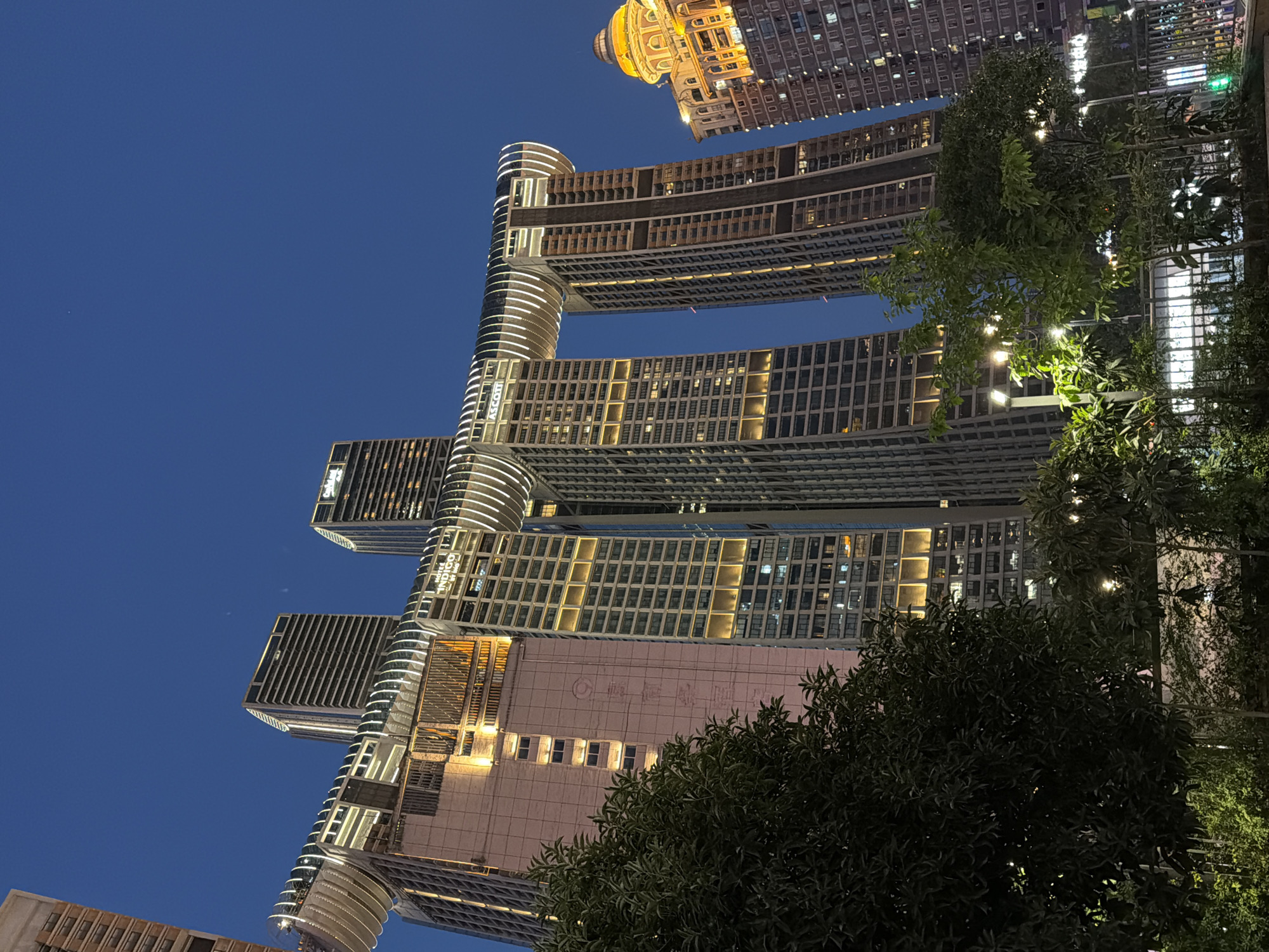 A low-angle, night shot captures the Raffles City Chongqing complex, a futuristic architectural marvel in the Yuzhong District of Chongqing, China. The complex, designed by Moshe Safdie, features four interconnected skyscrapers and a horizontal glass skybridge called "The Crystal" that spans across two of the towers, resembling a ship or a dragon.
The buildings are illuminated, with warm yellow lights accentuating the windows and vertical lines of the skyscrapers. The skybridge itself is outlined with bright white LED lights, creating a striking contrast against the deep blue twilight sky. The skybridge houses various amenities, including observation decks and restaurants, offering panoramic views of the city.
In the foreground, lush green trees and bushes are visible, partially obscuring the lower sections of the buildings and creating a sense of depth. The foliage is illuminated by soft, ambient lighting, adding to the tranquil nighttime atmosphere. In the distance, another building with a domed roof, illuminated in golden hues, adds to the vibrant cityscape. The image conveys a sense of modern urban development and architectural innovation within the bustling city of Chongqing.