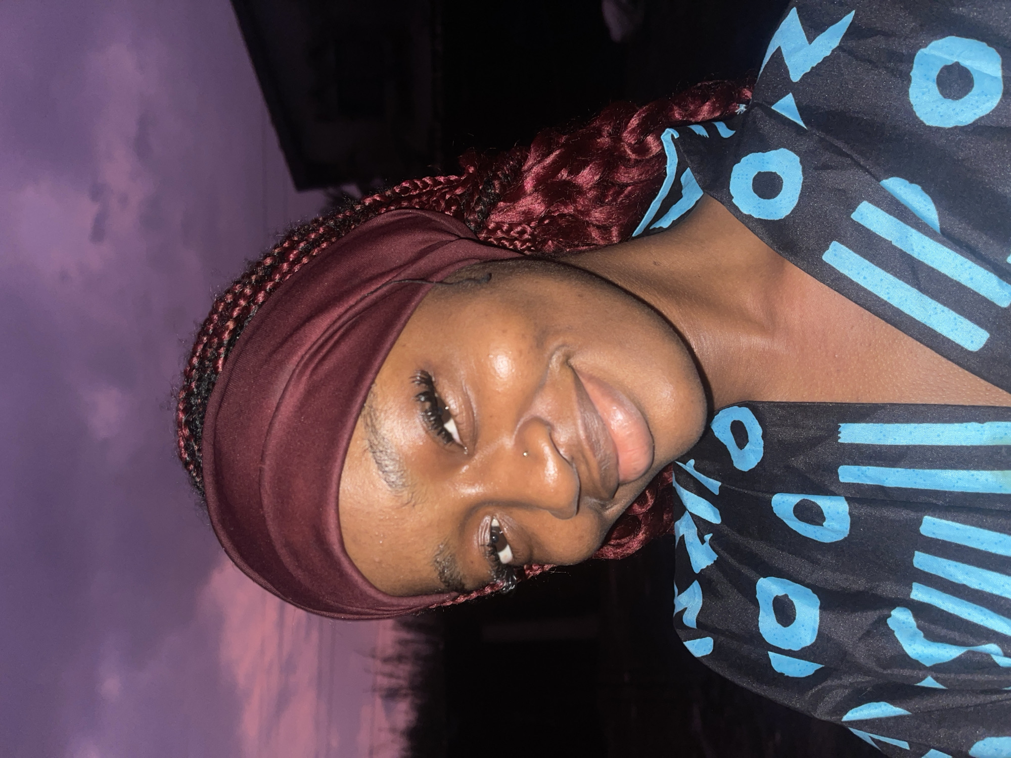 A medium shot portrait of a young Black woman is captured outdoors at dusk, with a dramatic purple sky filled with clouds in the background. She is facing the camera, her head tilted slightly to her left, with a warm smile. She is wearing a maroon headscarf that covers her hair, with some dark red braids visible peeking out from underneath. Her clothing is a black garment with a repeating pattern of light blue shapes, resembling abstract symbols or letters. A house structure is visible in the far right background, silhouetted against the twilight sky. The lighting suggests the photo was taken with a flash or in low-light conditions, highlighting the woman's features and giving her skin a radiant glow. Based on the context provided, this image is situated in Ado, Nigeria.