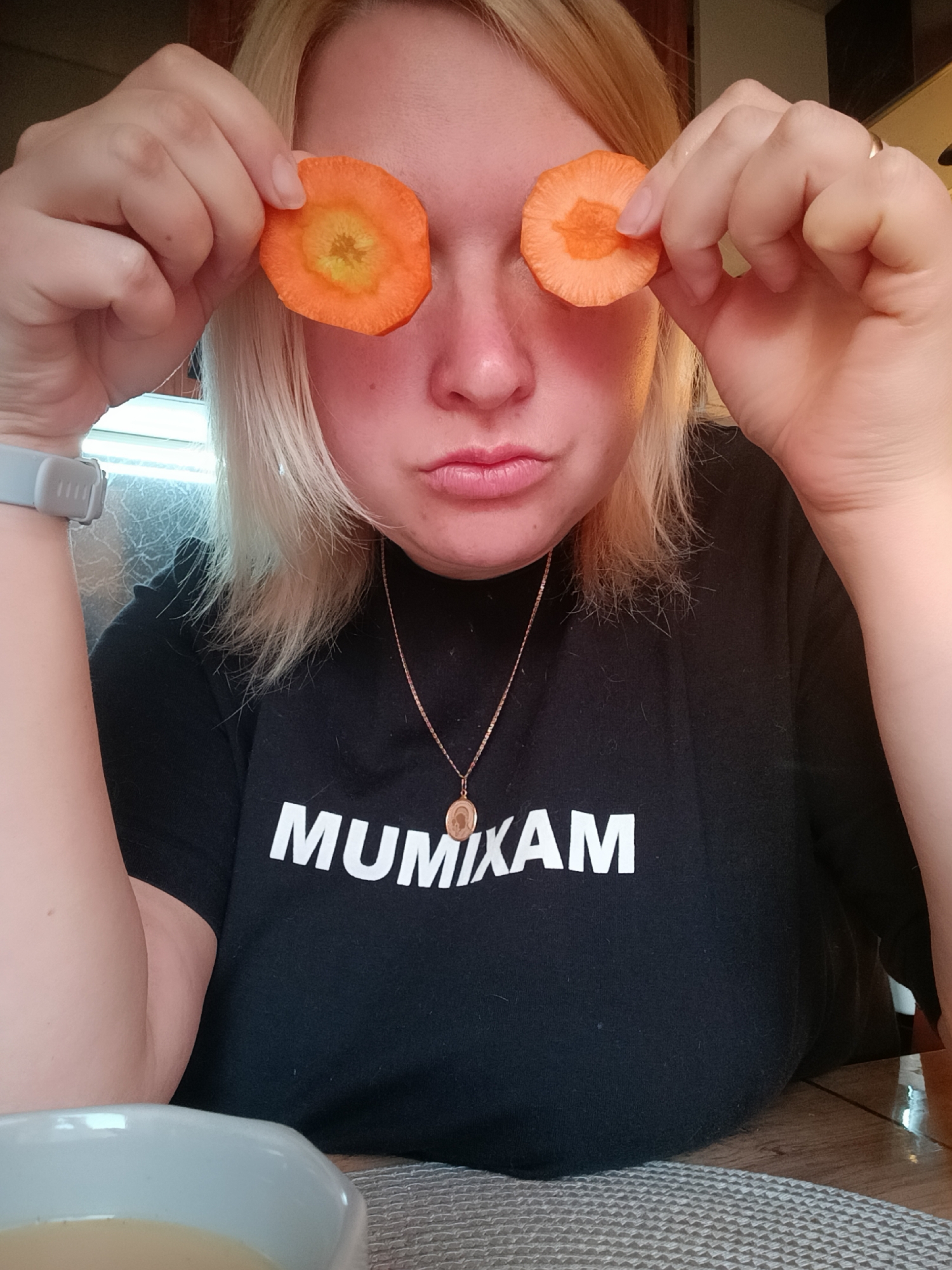 A woman with blonde hair is holding two slices of carrot over her eyes, obscuring them. She wears a black t-shirt with "MUMIXAM" in white letters. A gold necklace with a pendant hangs around her neck. She has fair skin and a slight pout. The woman wears a smartwatch on her left wrist. A bowl of light-colored liquid rests in the foreground. The table has a grey woven placemat. The background is slightly out of focus, but seems to be a kitchen setting. The image was likely taken indoors. The woman is not located in the city of Chaikovsky, Russia.