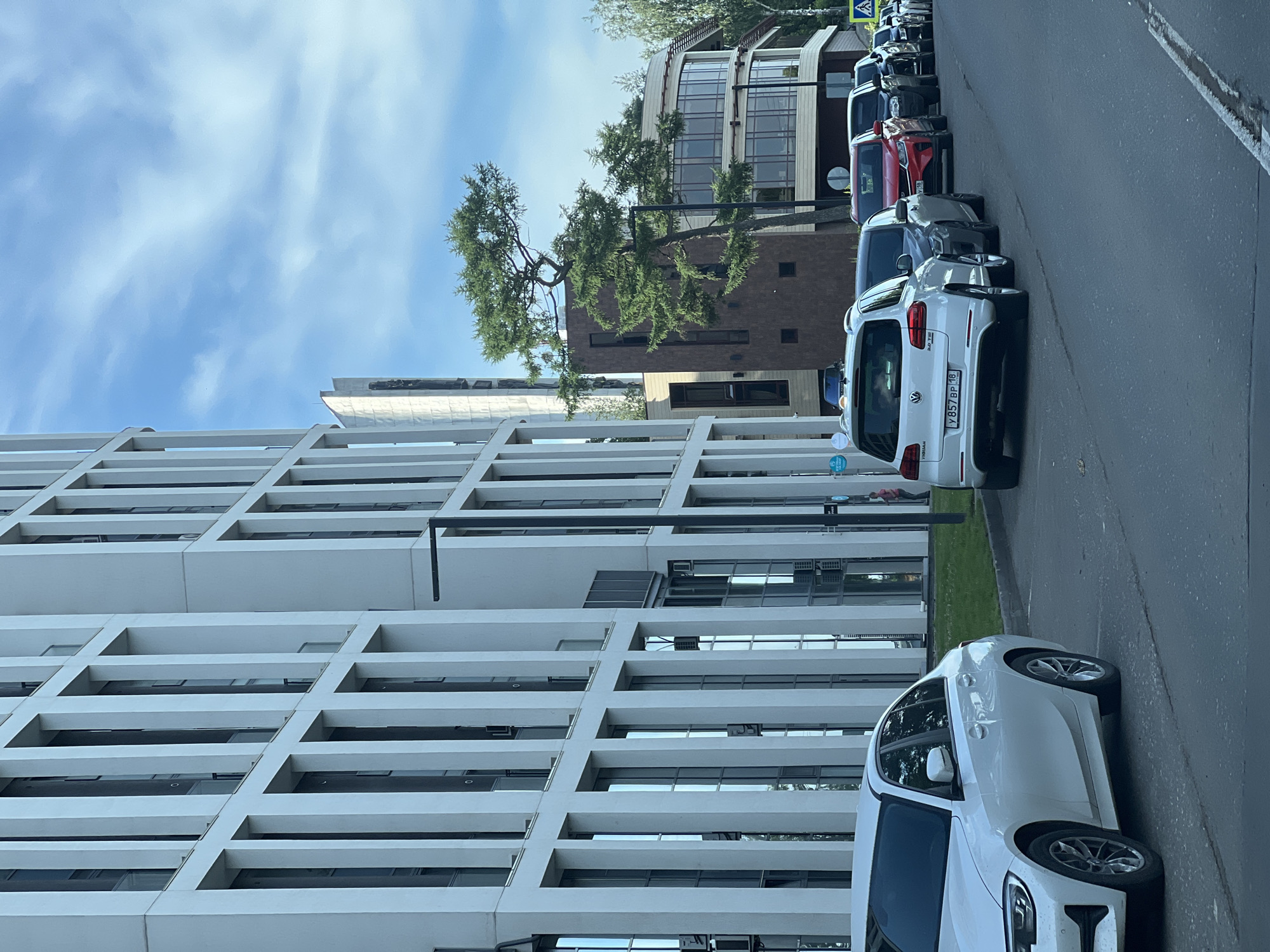 The image depicts a street scene in front of a modern, white, multi-story building with numerous vertical window sections. To the right of this building, a brown, multi-story structure with rounded glass-enclosed sections is visible, along with a tall, distinctive evergreen tree. A line of parked cars is present on the street. The foreground features the front of a white sedan. Further down the street, a white SUV with a Russian license plate (Y857BP 18) is parked, followed by other vehicles including a red sedan and several dark-colored cars. A pedestrian crossing sign is visible in the distance. The sky is partly cloudy and blue. This scene is consistent with an urban environment in Izhevsk, Russia, based on the license plate format and the architectural style.