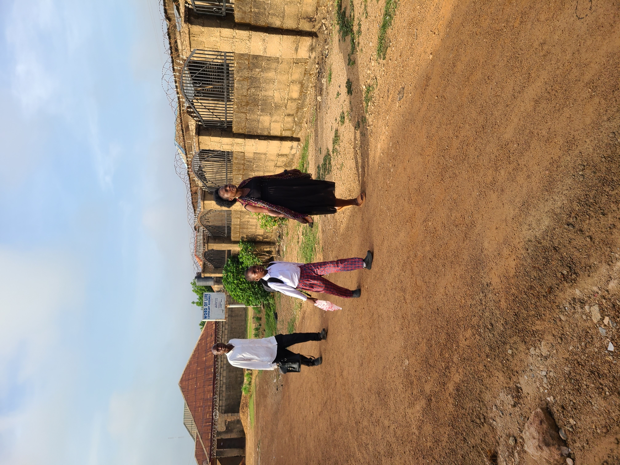 The image depicts an outdoor scene in Barakin Lerawi, Nigeria. Three people are walking along a reddish-brown dirt road. A woman wearing a dark dress with patterned trim walks closest to a brick wall topped with barbed wire. A young person in a white shirt and red-and-black plaid pants walks between her and a man who is wearing a white shirt and black pants. The man carries a black bag. Behind them is a building with a rusty roof and a sign that reads "WORD OF LIFE COVENANT ASSEMBLY". The sky is pale blue. The overall scene is bathed in sunlight.
