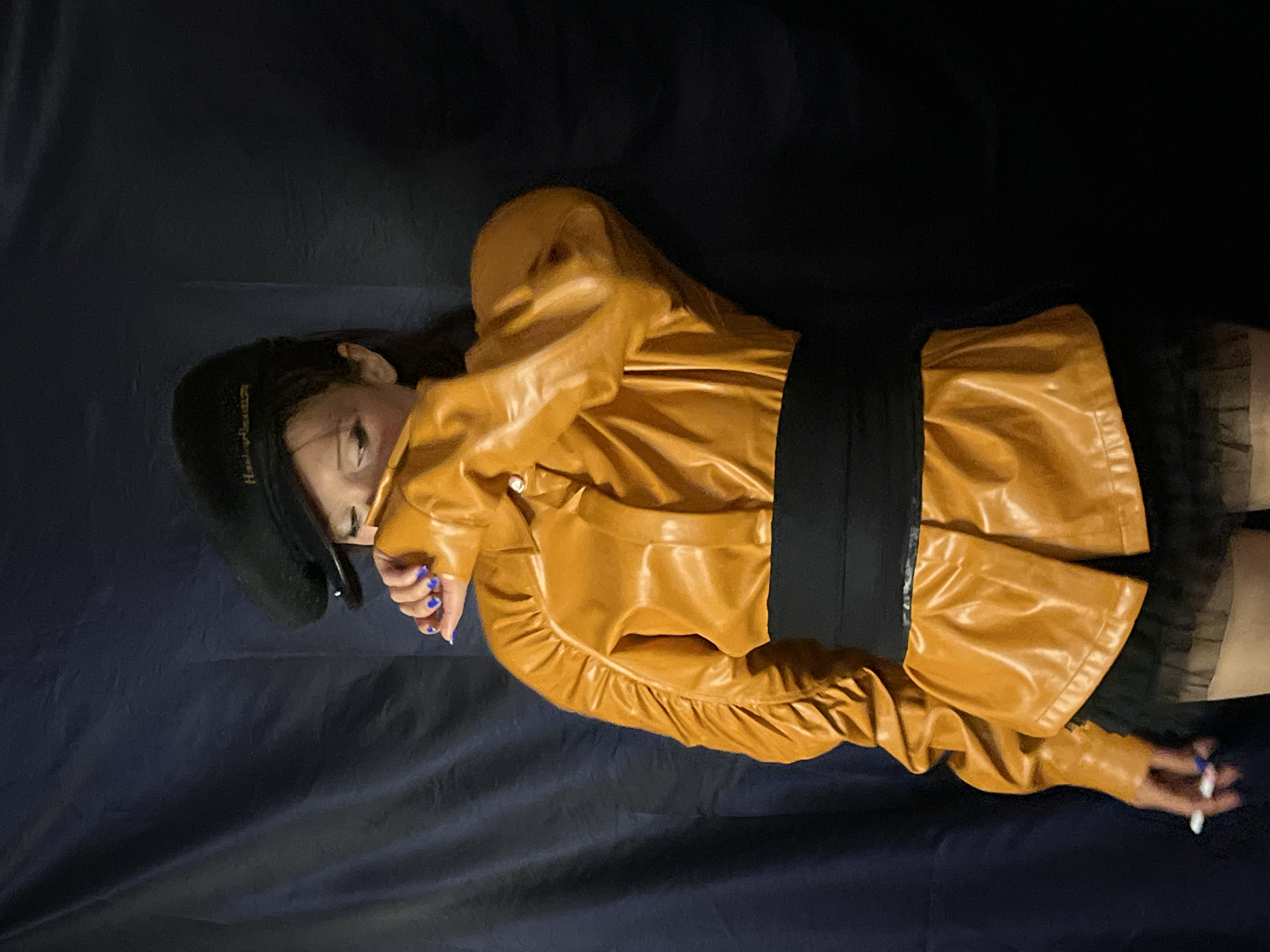 The image shows a person lying on a dark blue surface. The person wears a black beret, a mustard-colored leather jacket with a black wide belt, and a dark skirt with a sheer overlay. The person has blue fingernails and holds a thin white object (possibly a cigarette or pen) in their right hand. The location is not identifiable as Barcelona, Spain, from the provided visual information.