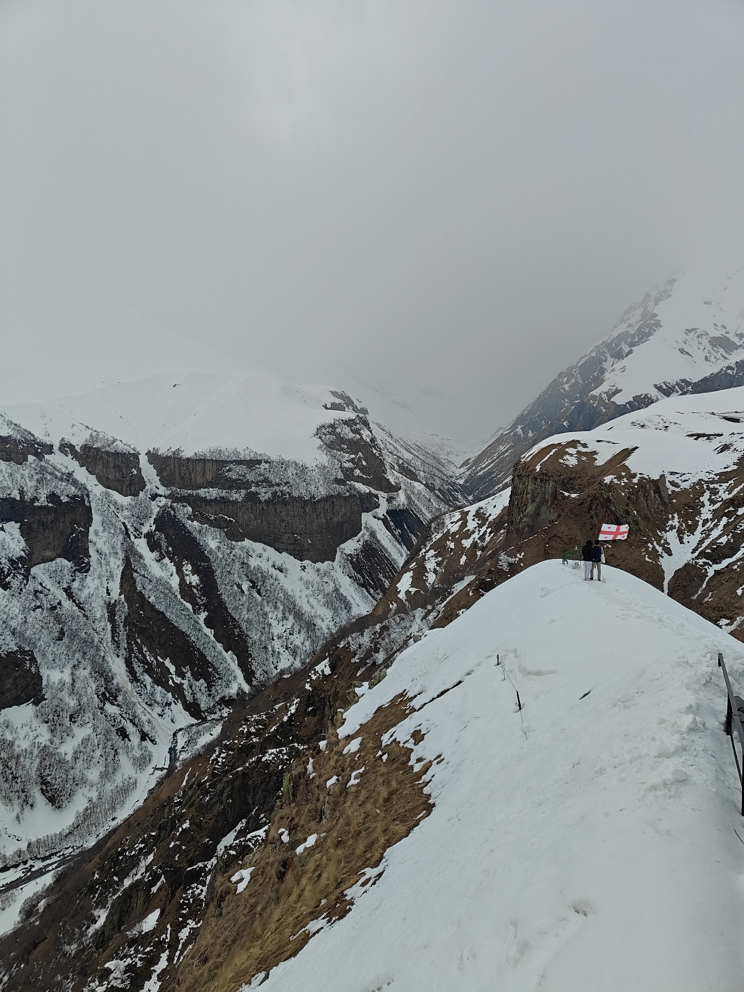 A wide shot captures a majestic, snow-covered mountain landscape under an overcast sky. In the mid-ground, two individuals stand on a narrow, snow-covered ridge, their backs to the camera, gazing out at the expansive valley and surrounding peaks. Behind them, a Georgian flag, white with five red crosses, is prominently displayed, affixed to a structure that is partially visible on the right.
The scene is dominated by rugged terrain, with steep, rocky cliffs and valleys blanketed in white snow. Darker rock faces and patches of sparse, snow-dusted trees contrast with the pristine snow. Some areas show patches of dry, brown grass peeking through the snow. A winding valley floor is visible in the distance, suggesting a river or road traversing it. The sky is a uniform light grey, indicating heavy cloud cover or fog, which partially obscures the distant mountain tops, lending an atmospheric and somewhat somber mood to the vast landscape. The presence of heavy snow suggests a winter or early spring setting in a high-altitude region.