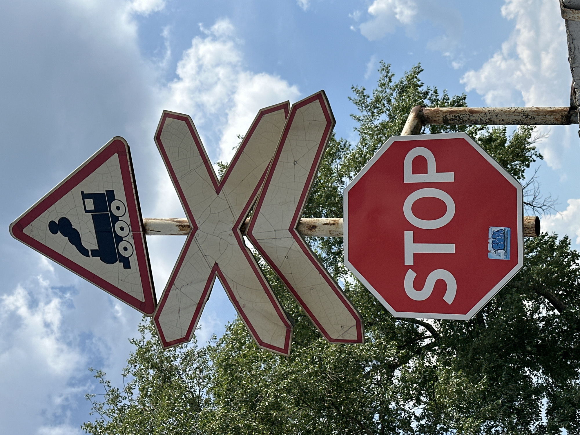 The image shows a traffic signpost with multiple signs in Selyatino, Russia. At the top, a triangular warning sign depicts a black steam train emitting smoke. Below this, there are three diagonal white directional arrow signs with red borders, arranged in a cross shape. The lowest sign is a red octagonal STOP sign with white lettering. A small sticker, possibly graffiti, is visible on the lower part of the STOP sign. The signs are mounted on a rusty metal pole. In the background, lush green foliage from a tree is visible on the left, and a blue sky with scattered white clouds is present above. The overall scene suggests an outdoor location, likely a road intersection with a railway crossing.